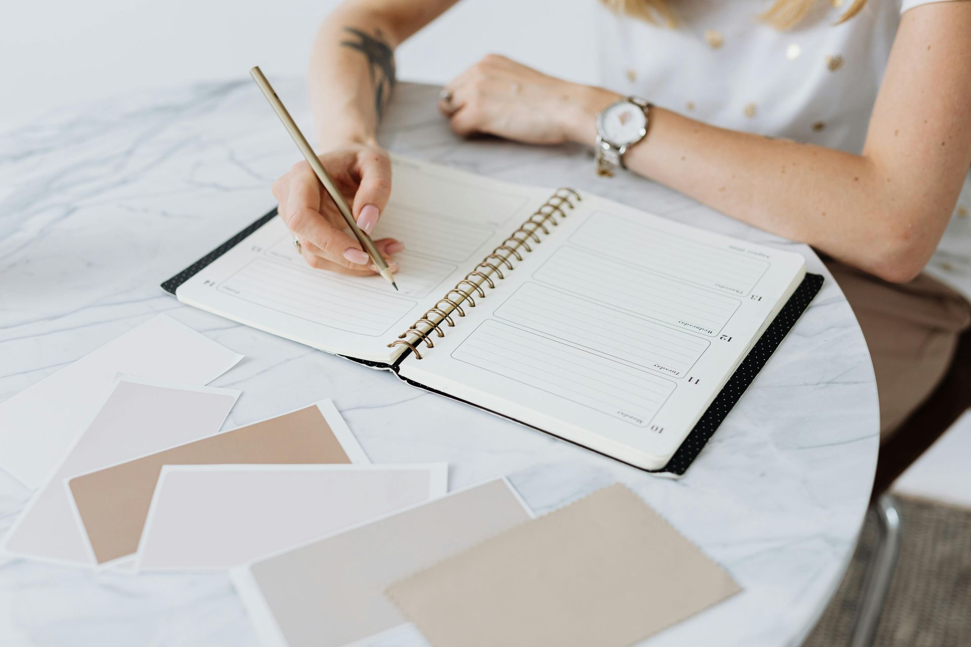 Person writing in a notebook at a marble table. Several color swatches are scattered around.