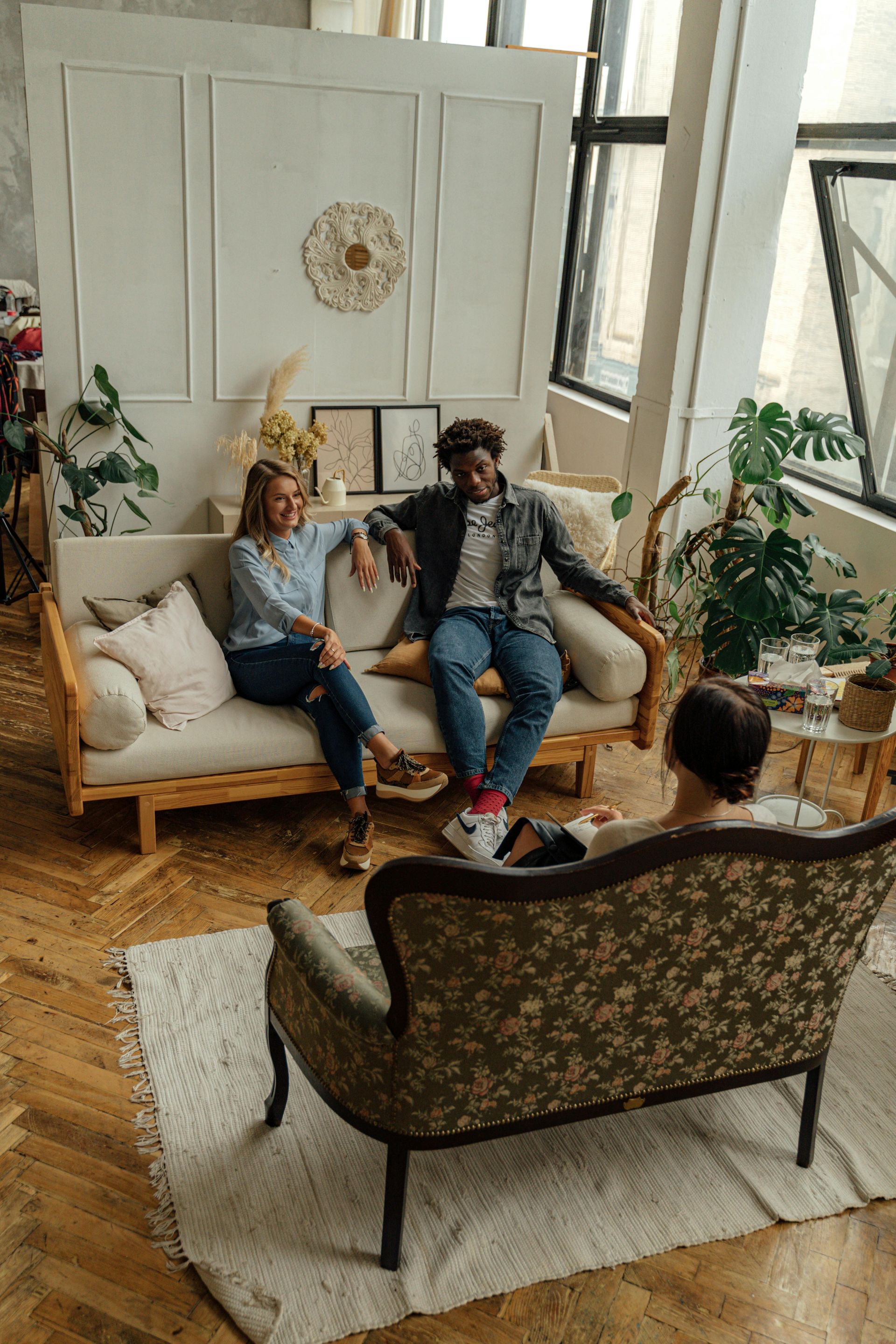 Three people conversing in a bright living room. One couple on a beige couch, another in a patterned chair.