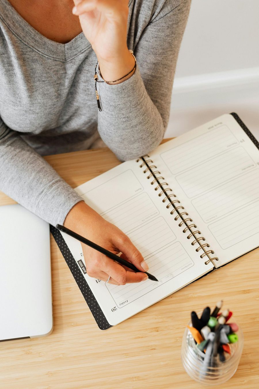 Woman writing in open notebook with a pen at a desk; next to a laptop, pen holder.