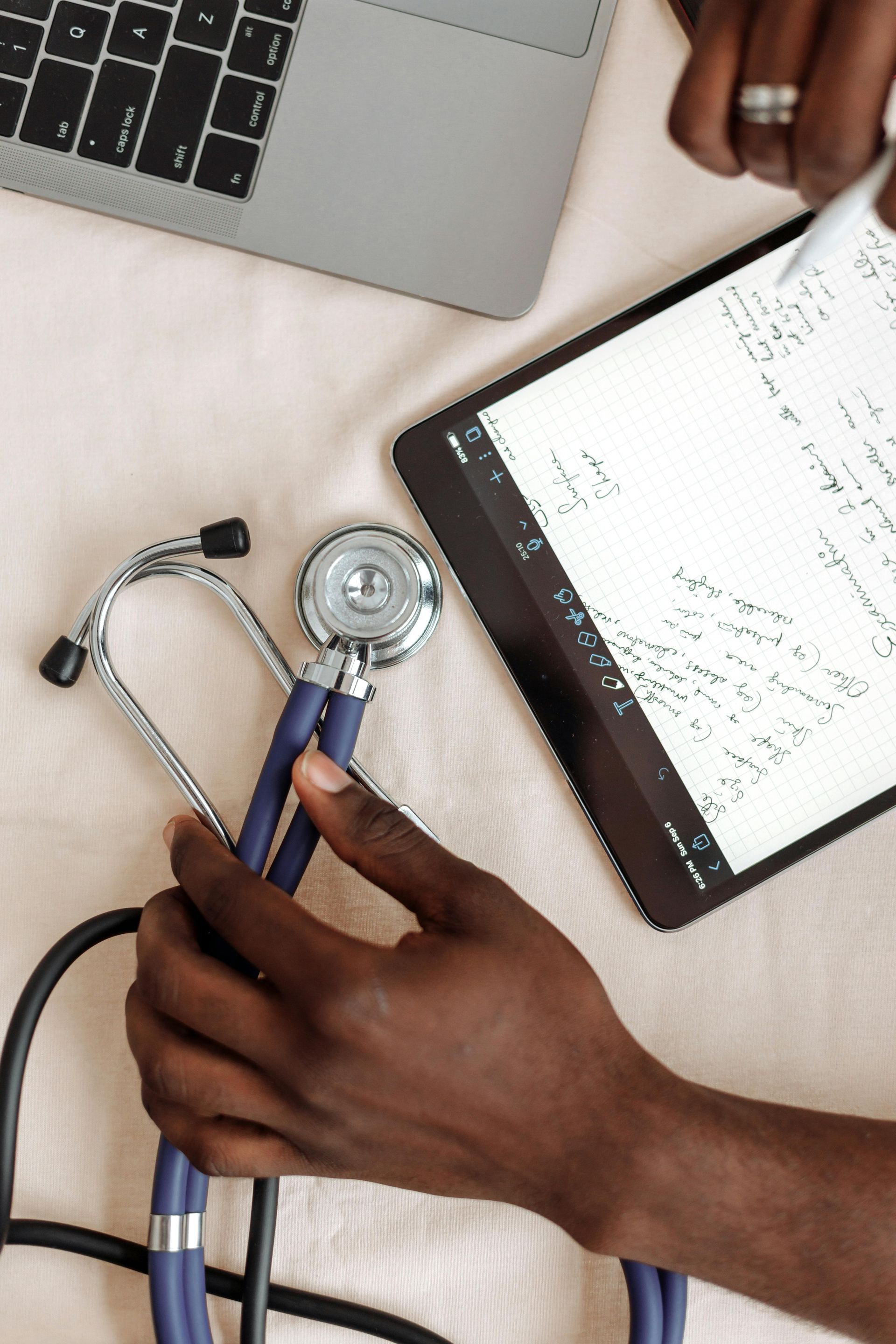 Person holding stethoscope, using tablet and laptop; medical setting.