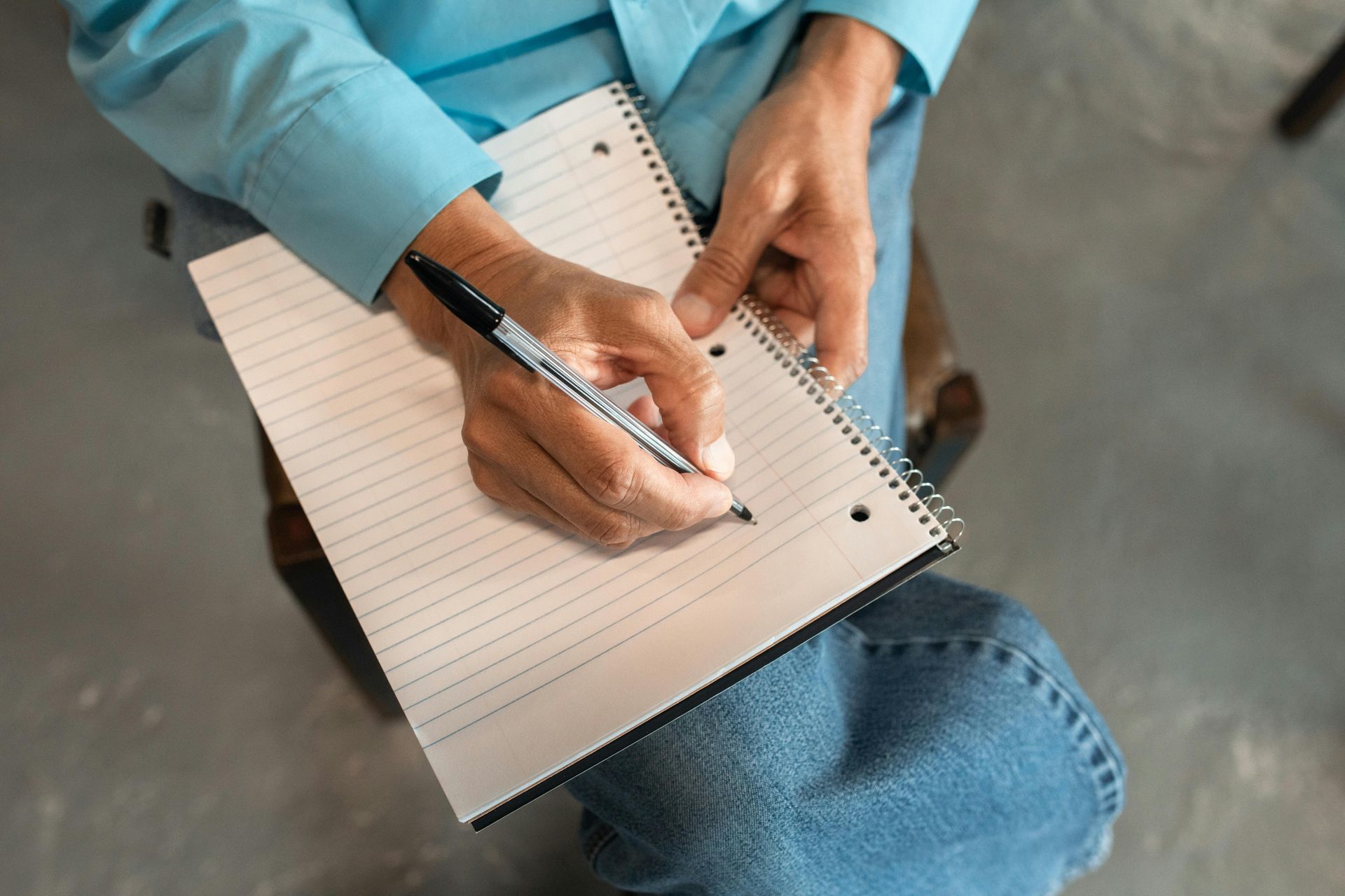 Person in blue shirt and jeans writing on a lined notepad with a black pen, seated indoors.