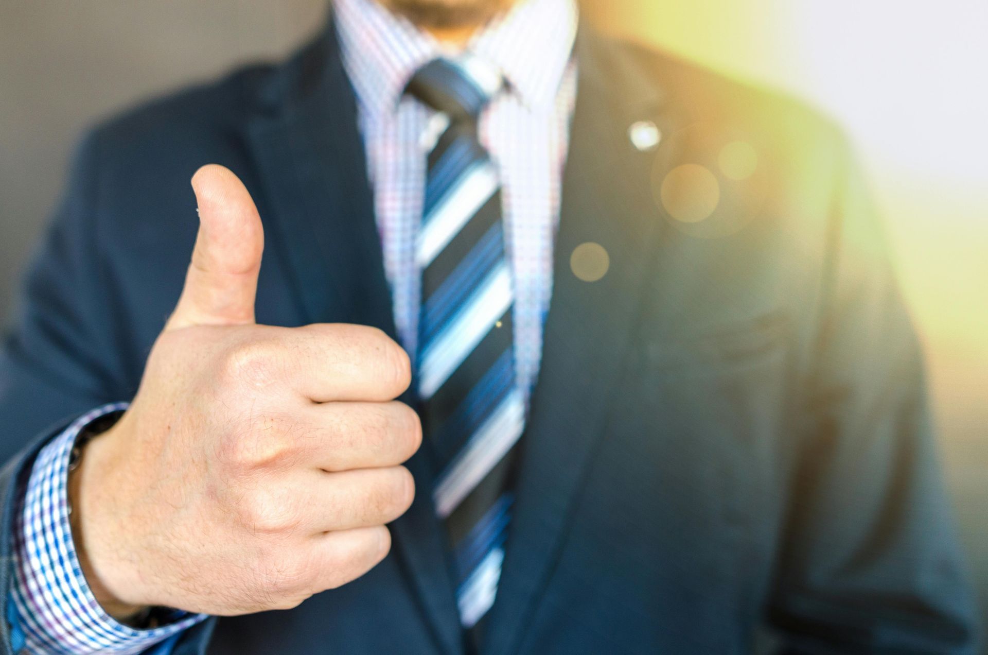 Man in a suit giving a thumbs-up. Blue striped tie, white and blue striped shirt. Sunlight on right.