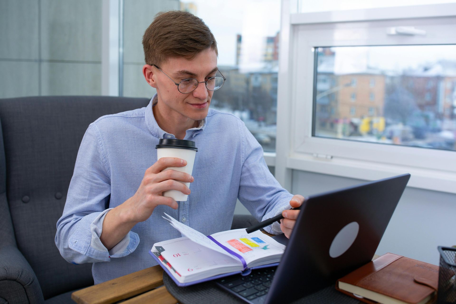 Man with glasses working on laptop, holding coffee, and looking at a notebook with charts.