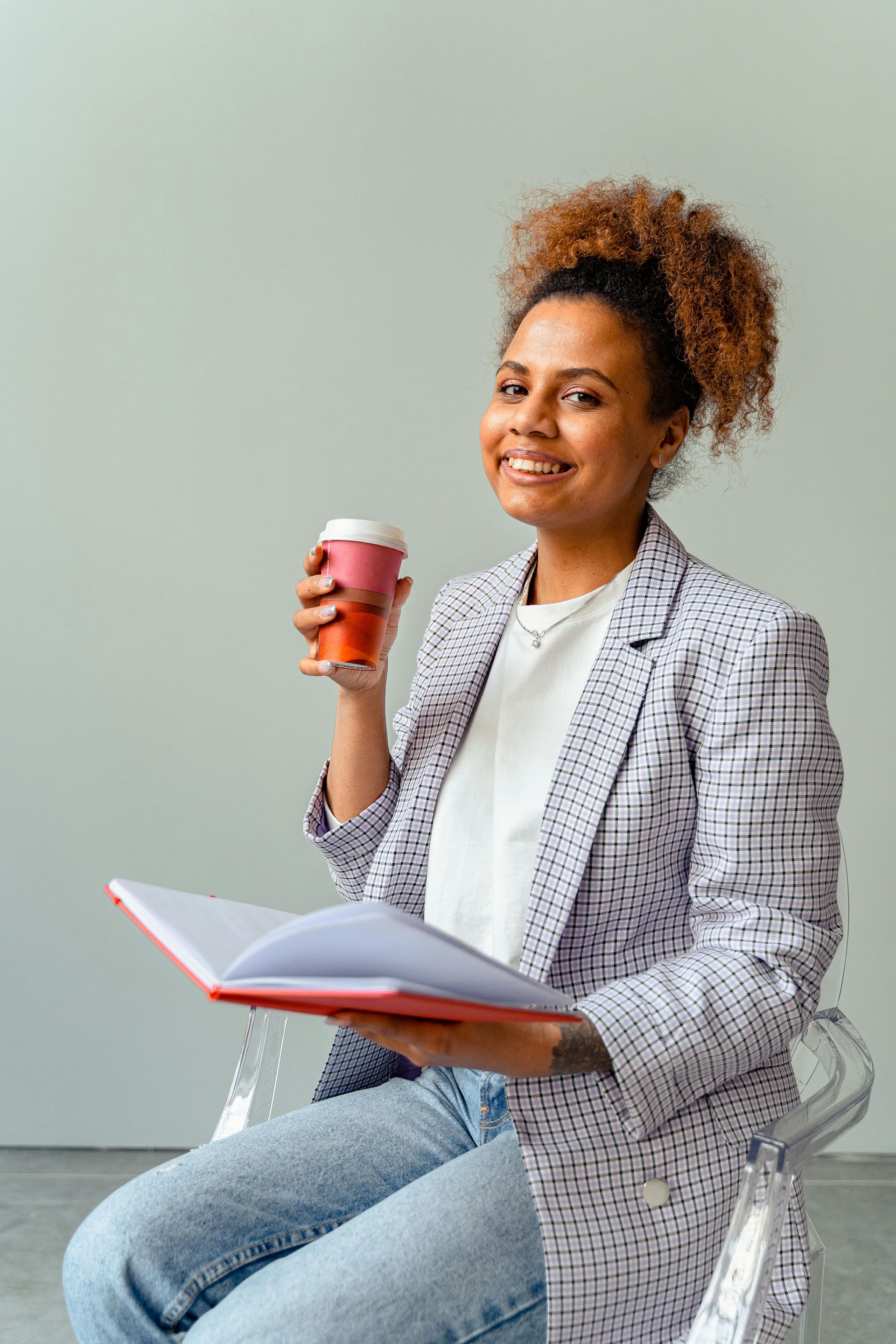 Woman holding coffee and notebook, smiling in a blazer, sitting in a clear chair.