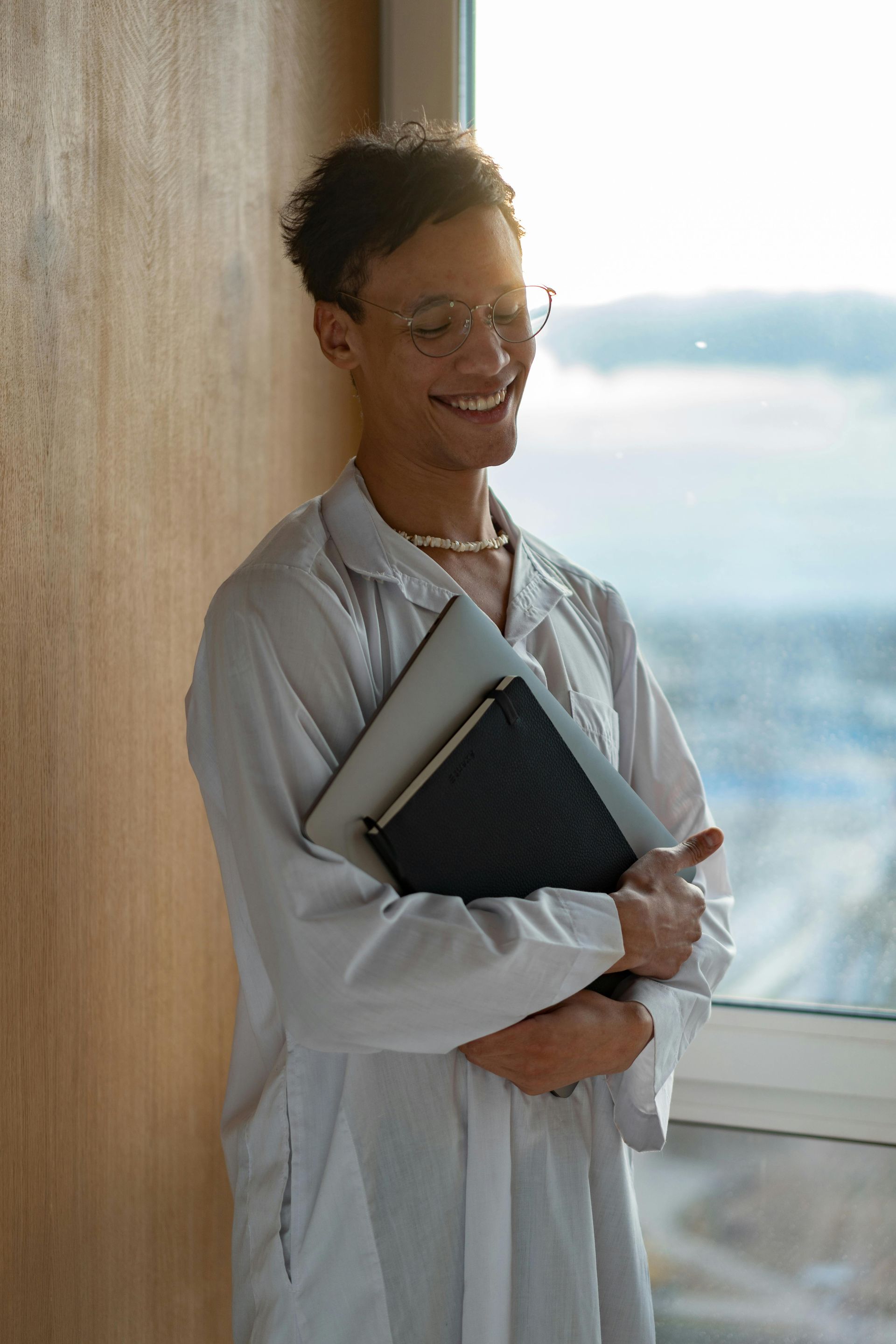 Smiling person in glasses and lab coat holding books by a window.