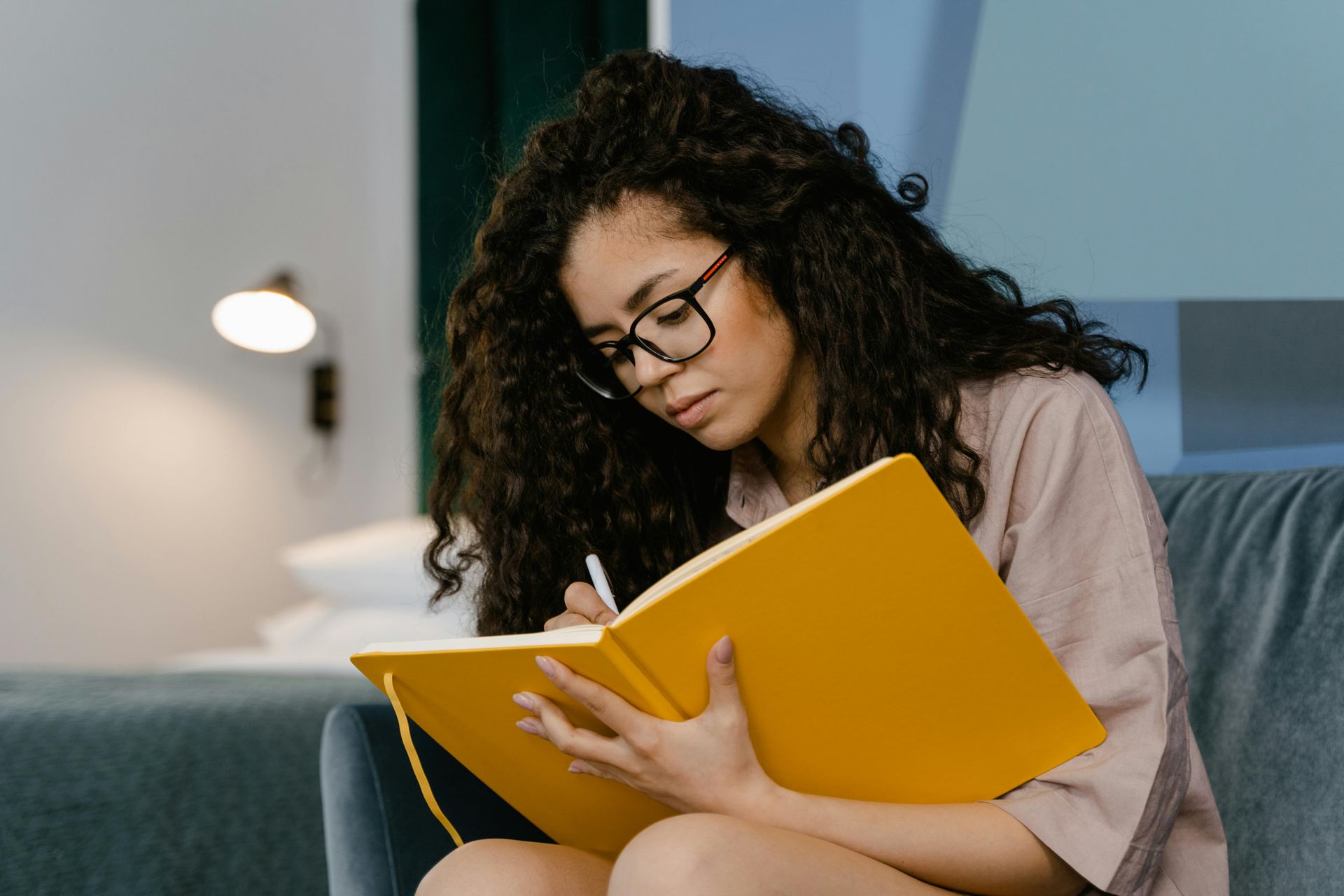 Woman with curly hair and glasses writes in a yellow notebook on a couch.