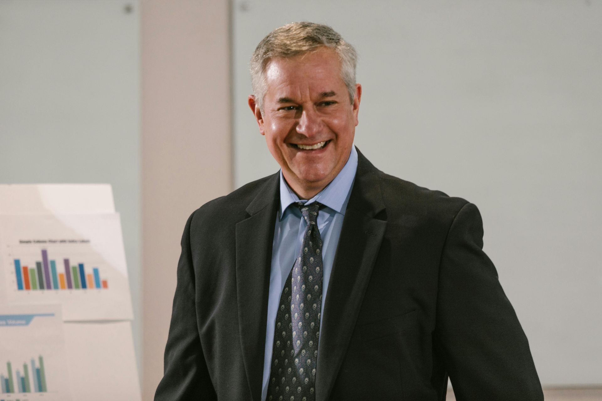 Smiling businessman in a dark suit, standing in front of a chart in a business setting.