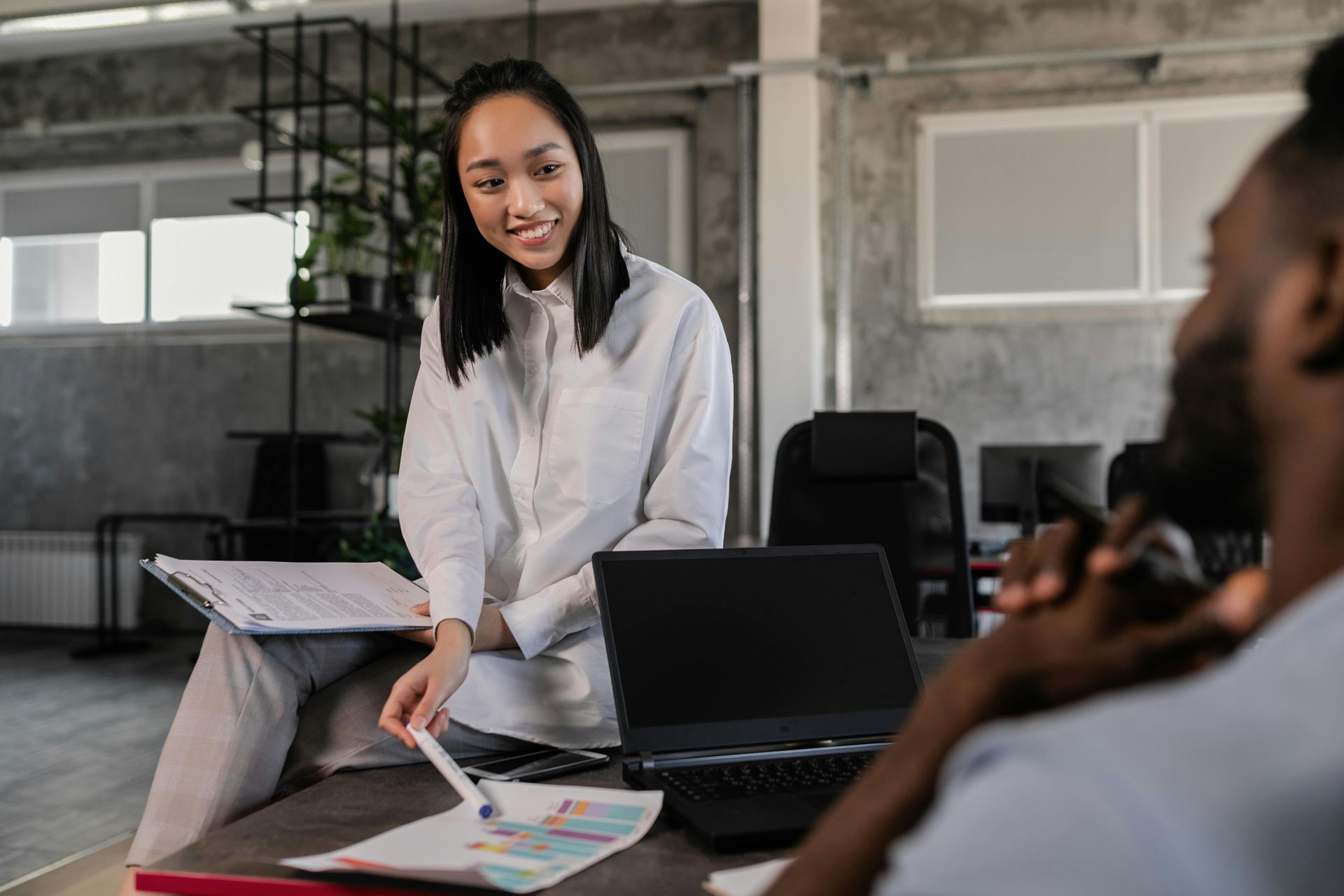 Woman sitting, smiling, pointing at documents on desk with Black man in office.