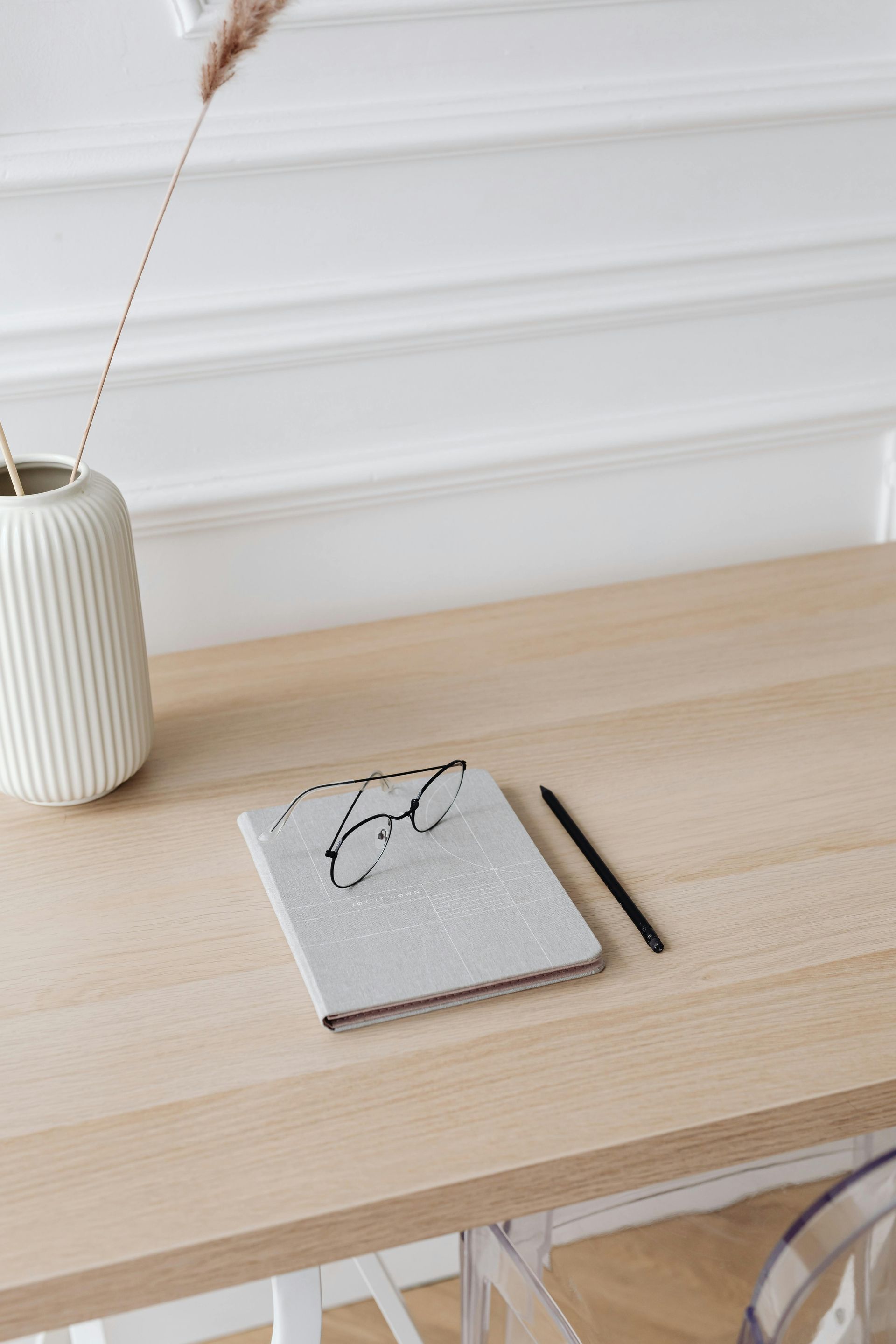 Notebook and pen on a light wood desk with a vase of dried plants.