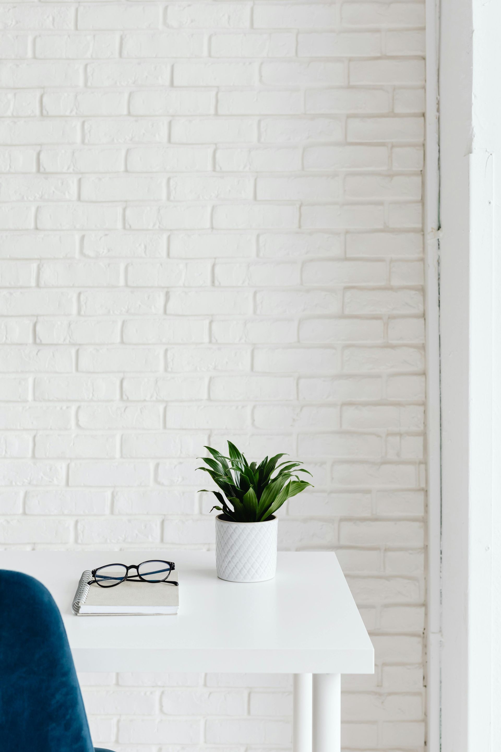 White desk with a potted plant, book, and glasses against a white brick wall; blue chair visible.