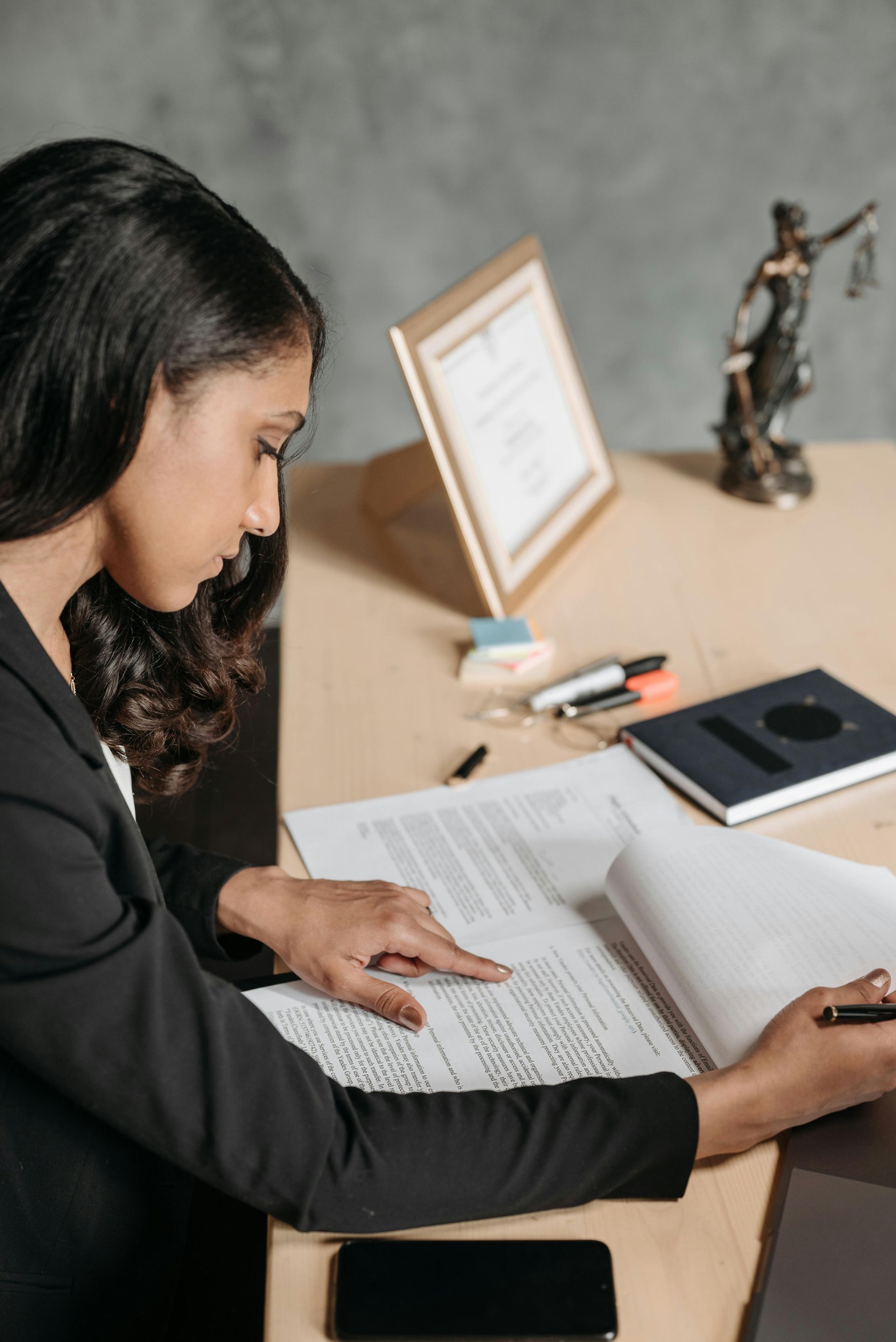 Woman in a black blazer reviews documents at a desk. A statue of Lady Justice and a framed certificate are nearby.