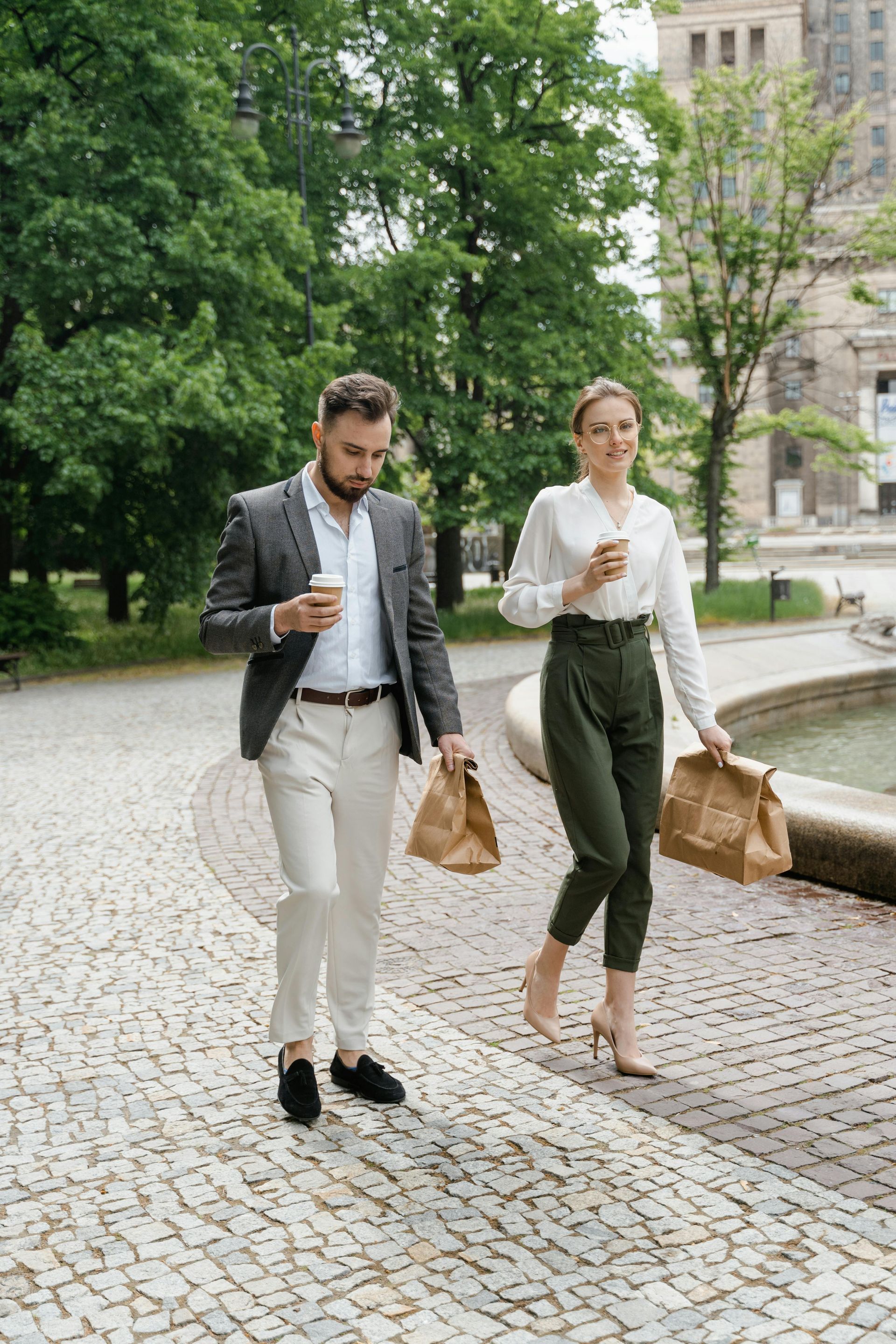 Man and woman walking, holding coffee and bags. He in blazer and slacks, she in white blouse and green pants. Park setting.