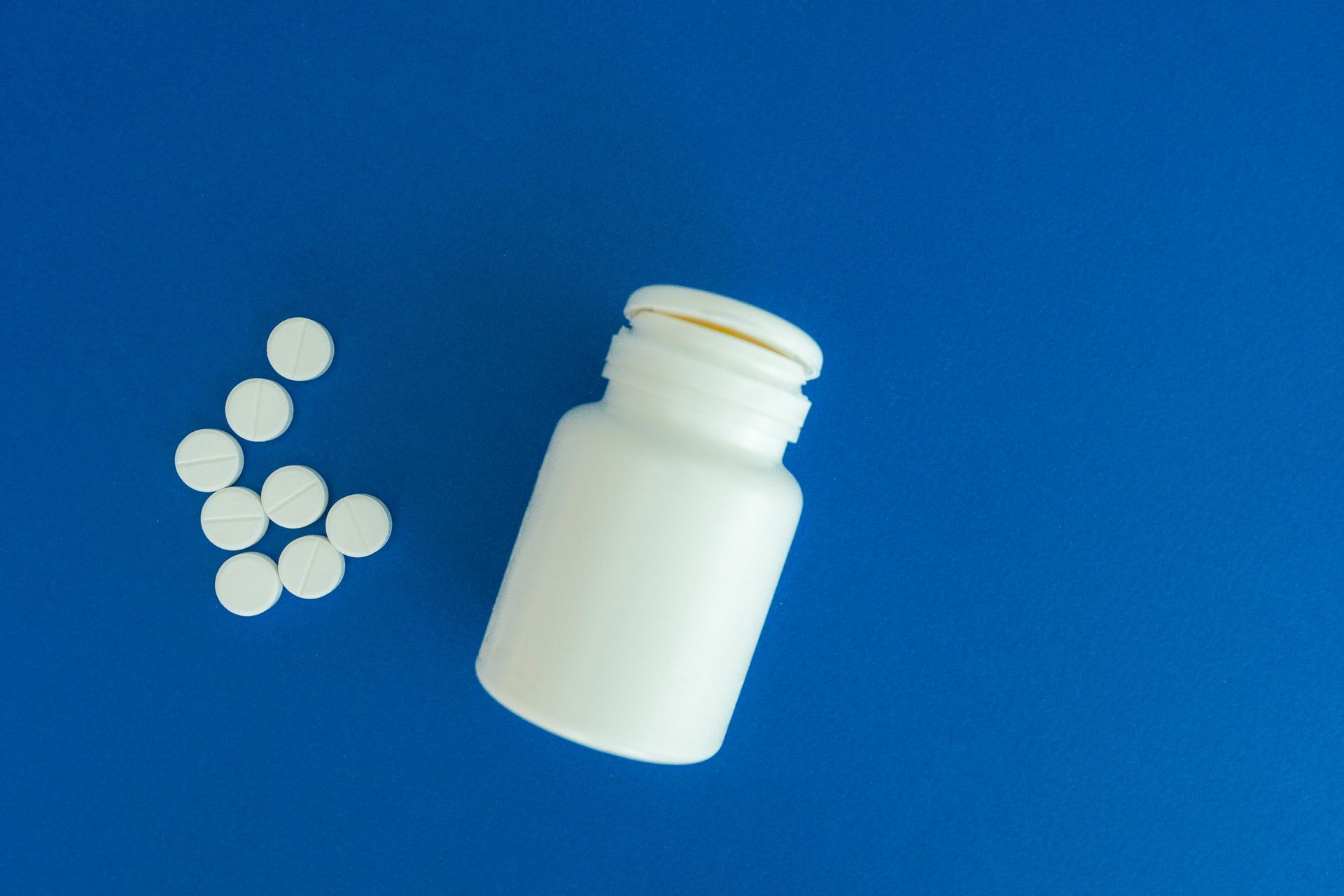 White pills next to a white pill bottle on a blue surface.