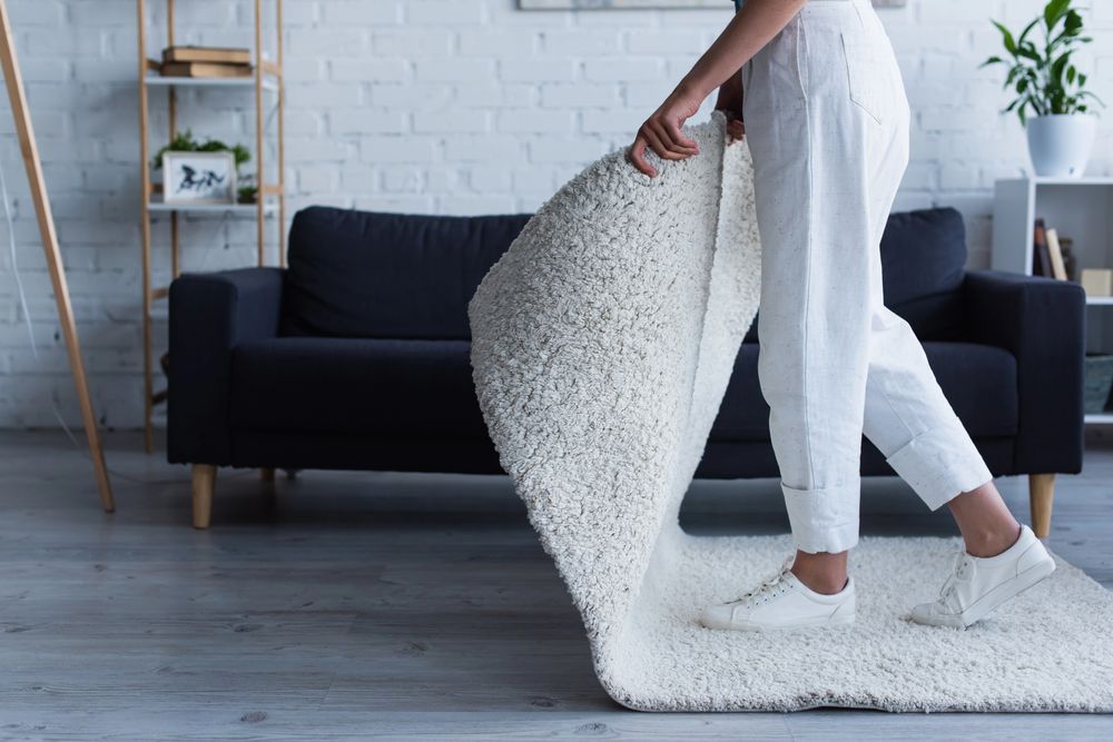 Persona vestida de blanco extiende una alfombra mullida de color crema en una sala de estar con un sofá negro.