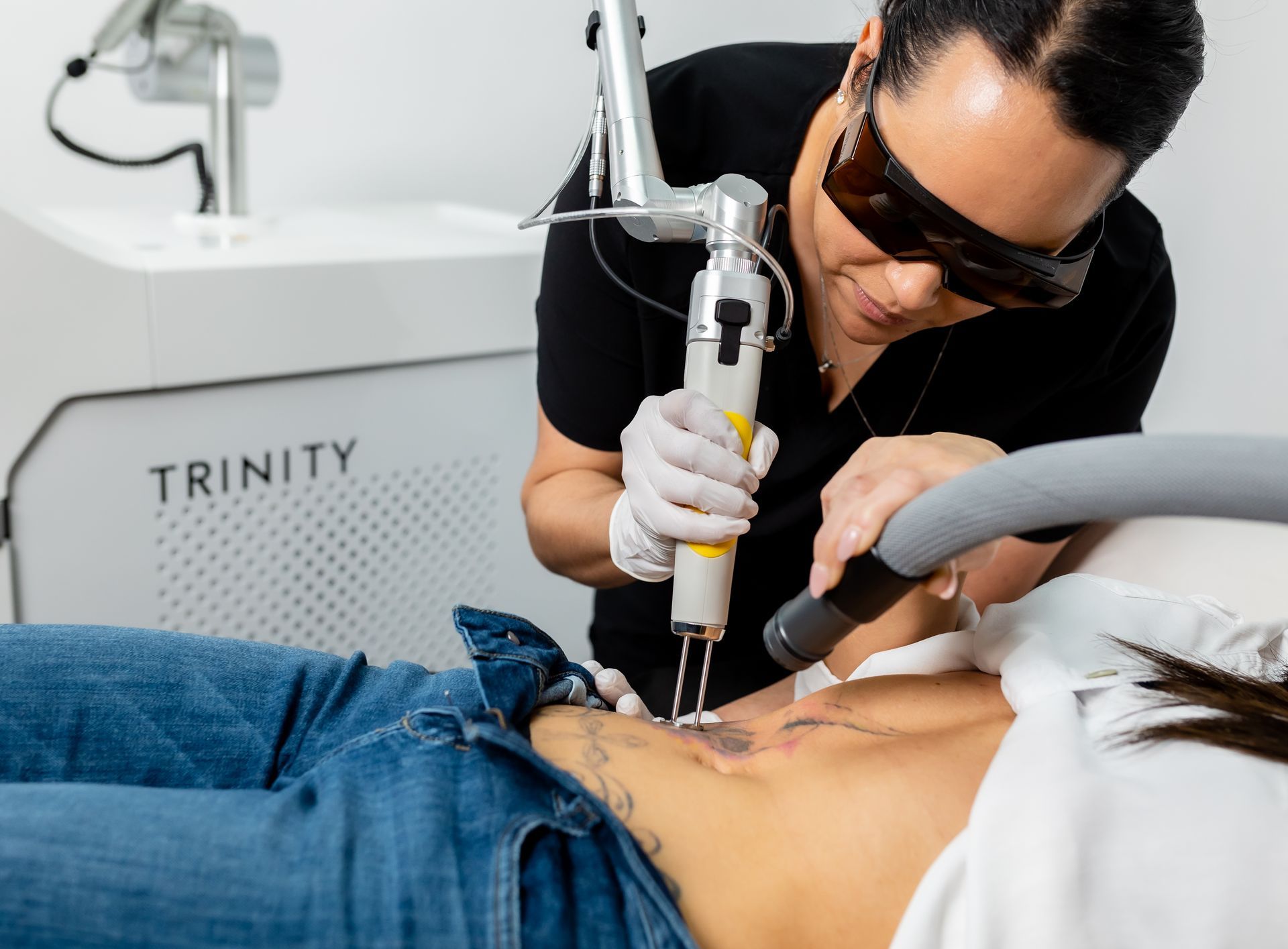 Medical professional using laser on a patient's abdomen, removing a tattoo. The setting is a clinic.