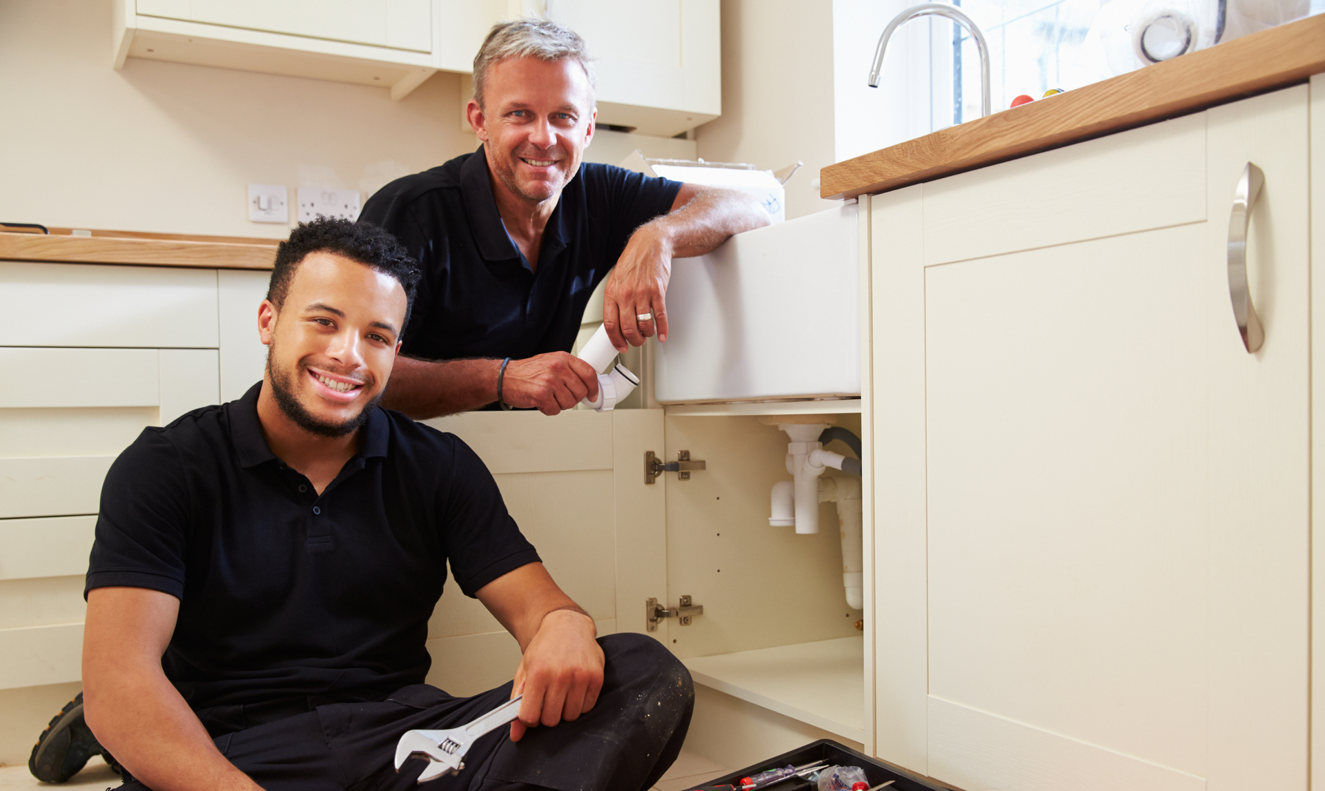 Two men are sitting on the floor in a kitchen fixing a sink.