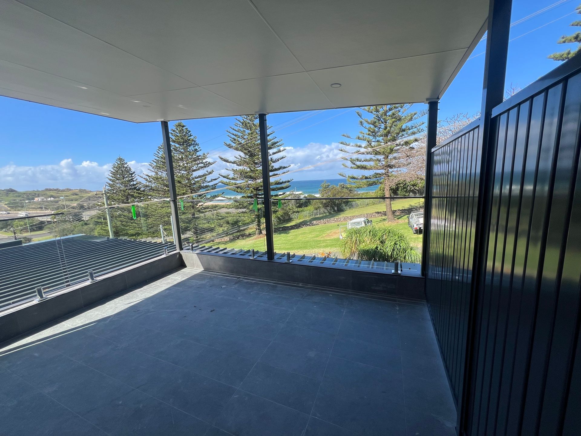 Covered patio with ocean view, black railing, and blue sky beyond a grassy landscape  — Taddeo Constructions in Kiama Downs, NSW