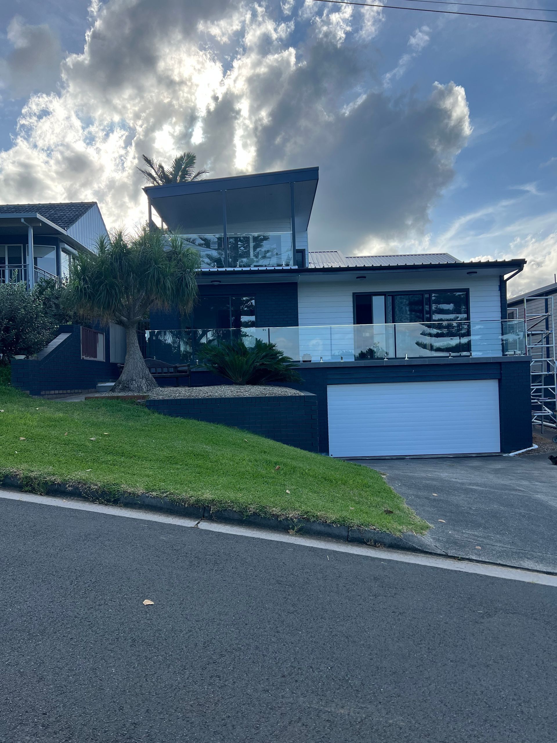 Modern house on a steep street under a cloudy sky, with a white garage door and green lawn  — Taddeo Constructions in Kiama Downs, NSW