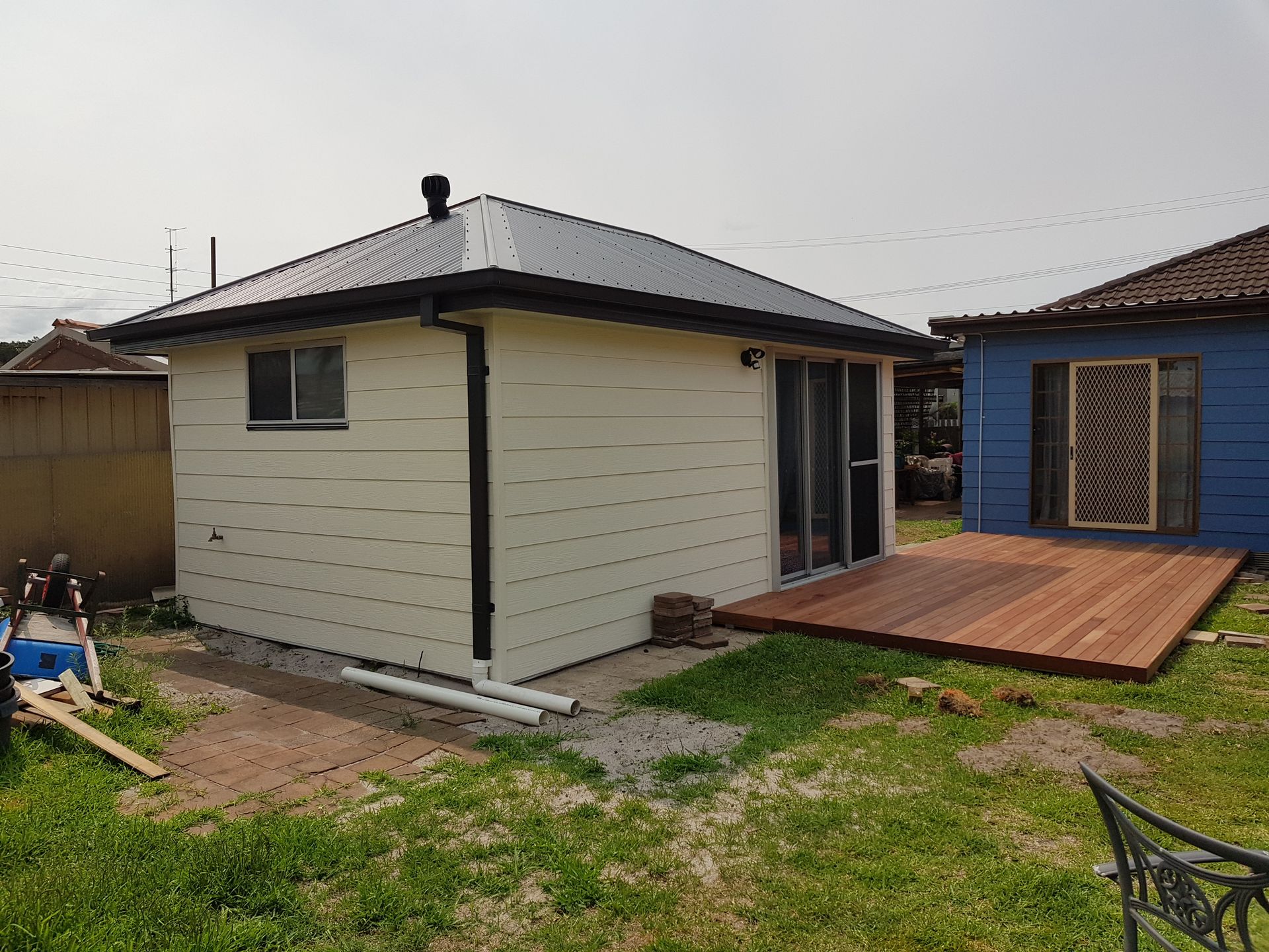 Small beige shed with dark roof and wooden deck in a grassy backyard, beside a blue garage. — Taddeo Constructions in Kiama Downs, NSW