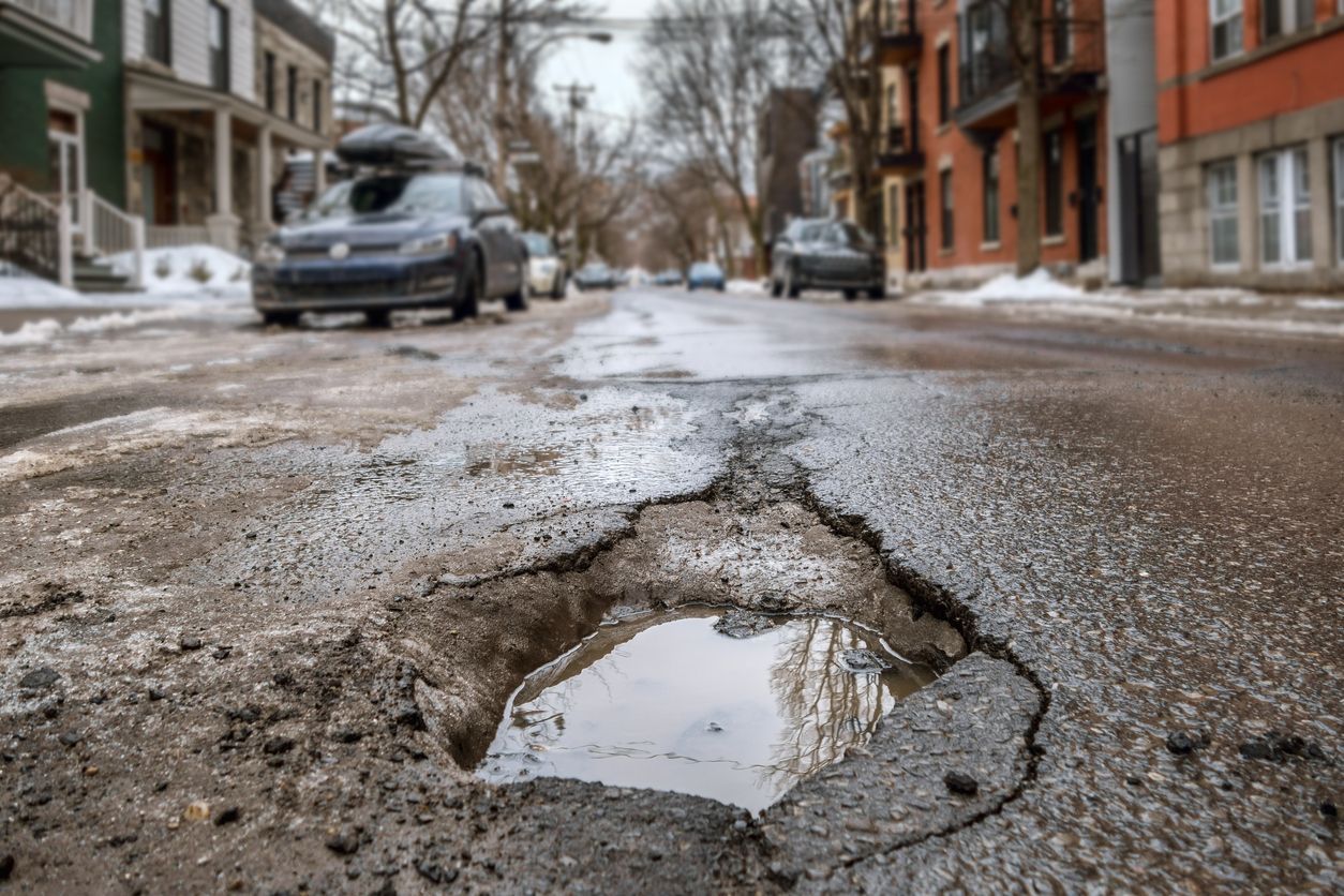 Low-pan view of a street with parked cars. In the middle is a large pothole full of water.
