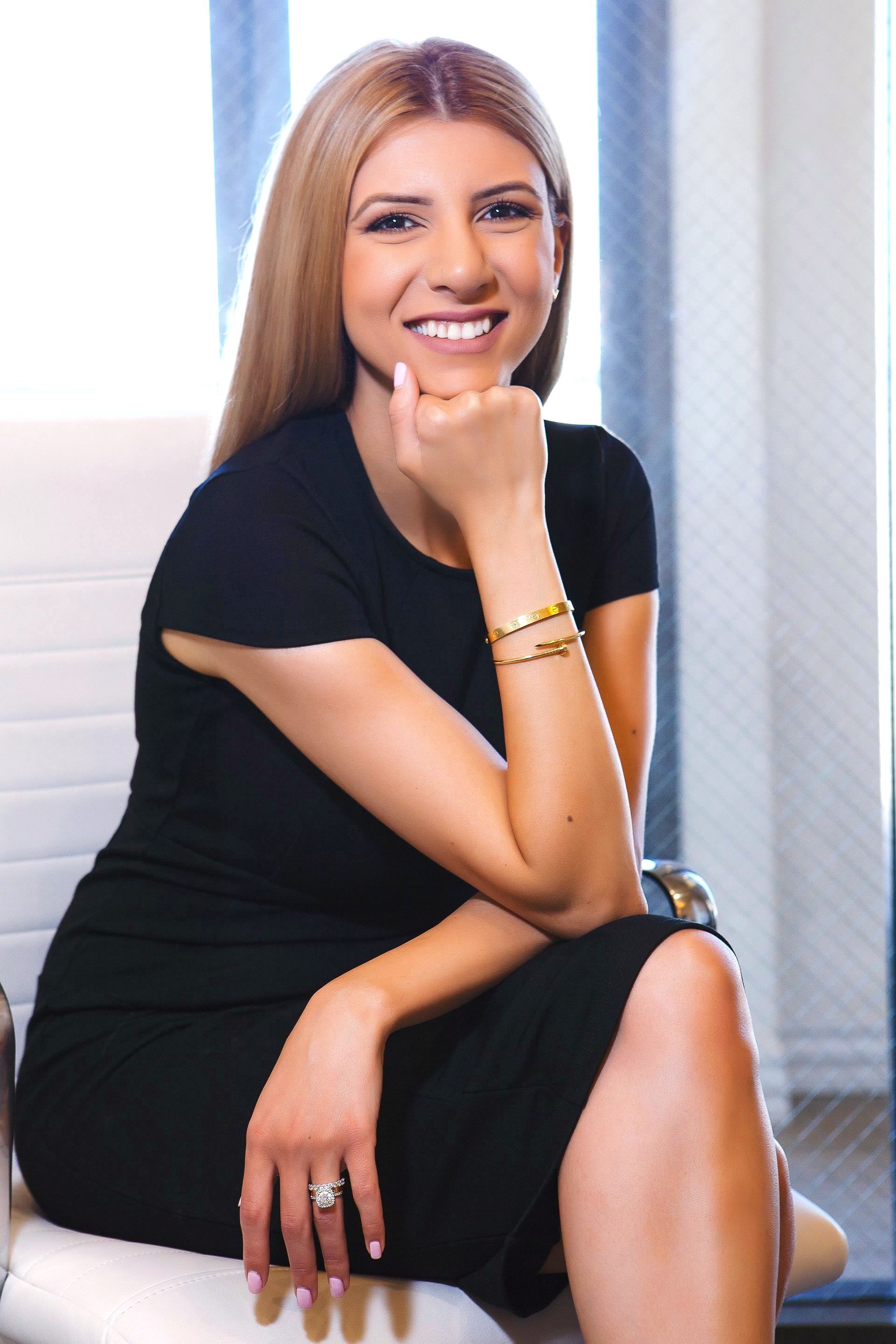 A woman in a black dress is sitting in a chair with her hand on her chin.