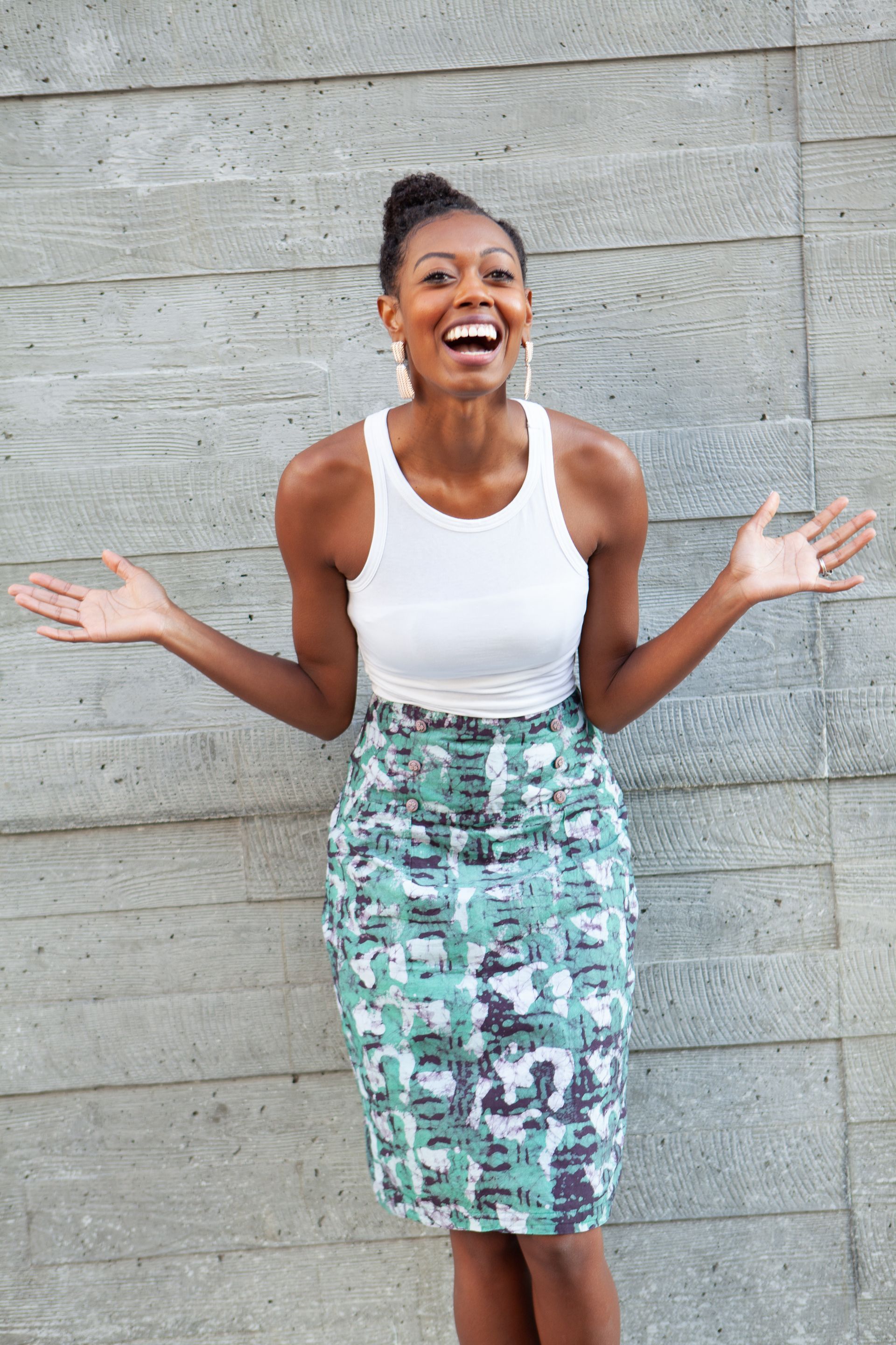A woman in a white tank top and a green skirt is standing in front of a concrete wall.