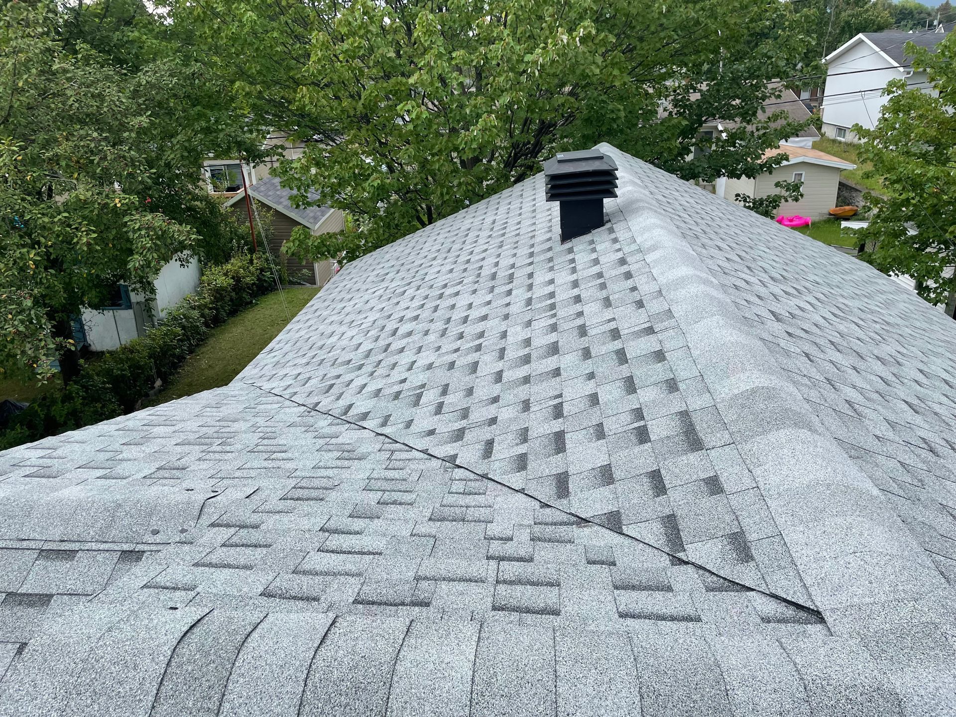 Toit en bardeaux gris avec une cheminée noire, entouré d'arbres verts et de maisons.