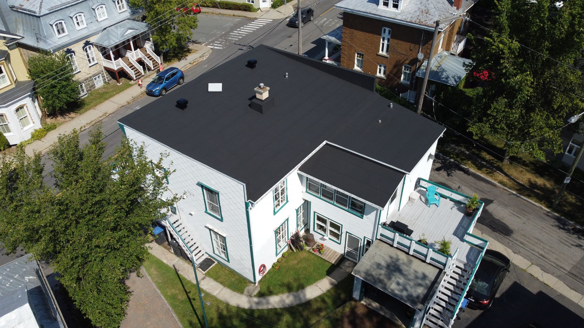 Vue aérienne d'un bâtiment blanc avec un toit noir, une terrasse et une rue environnante avec des arbres et d'autres bâtiments.