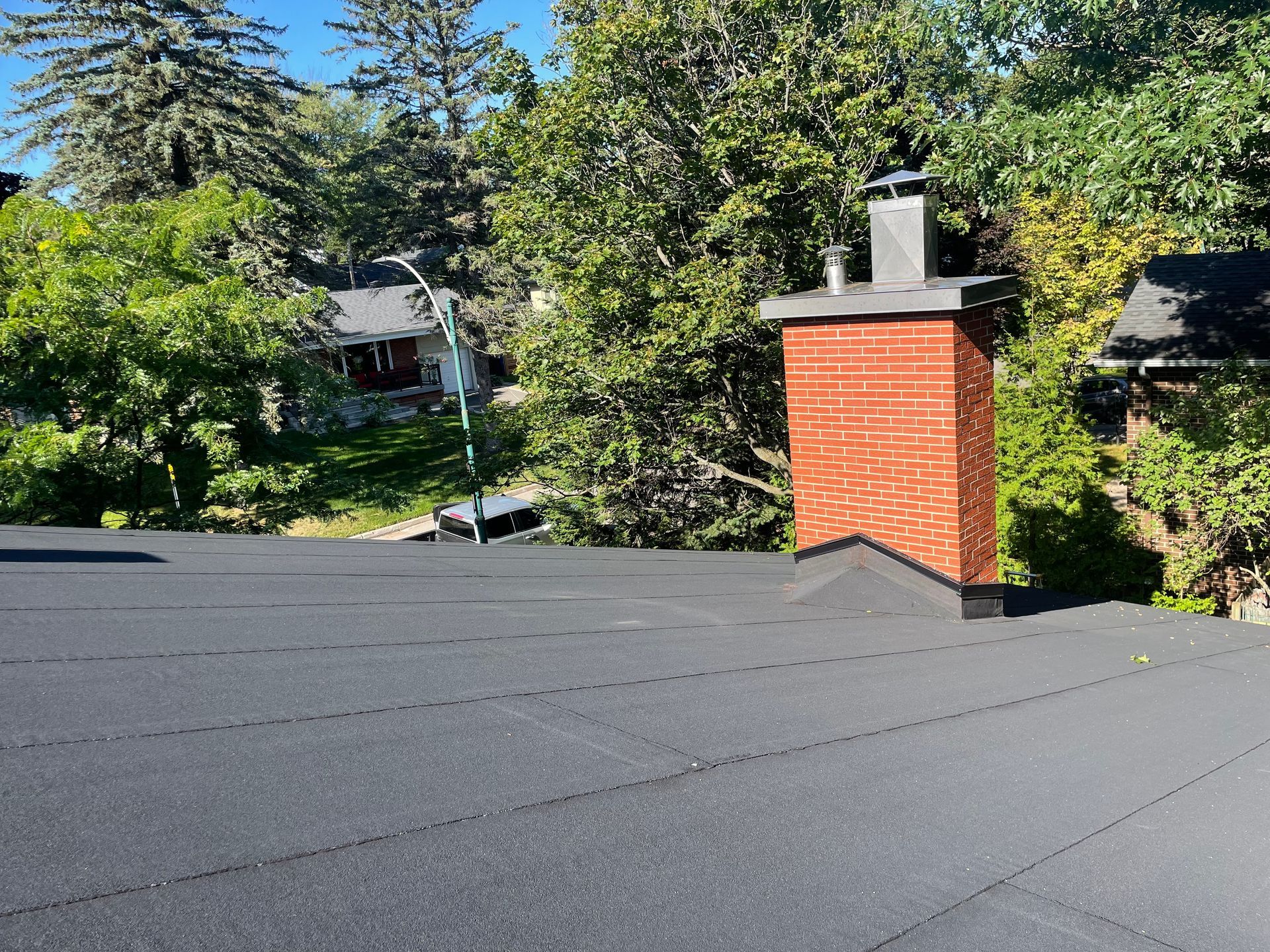 Toit plat noir avec cheminée en briques entourée d'arbres verts par une journée ensoleillée.