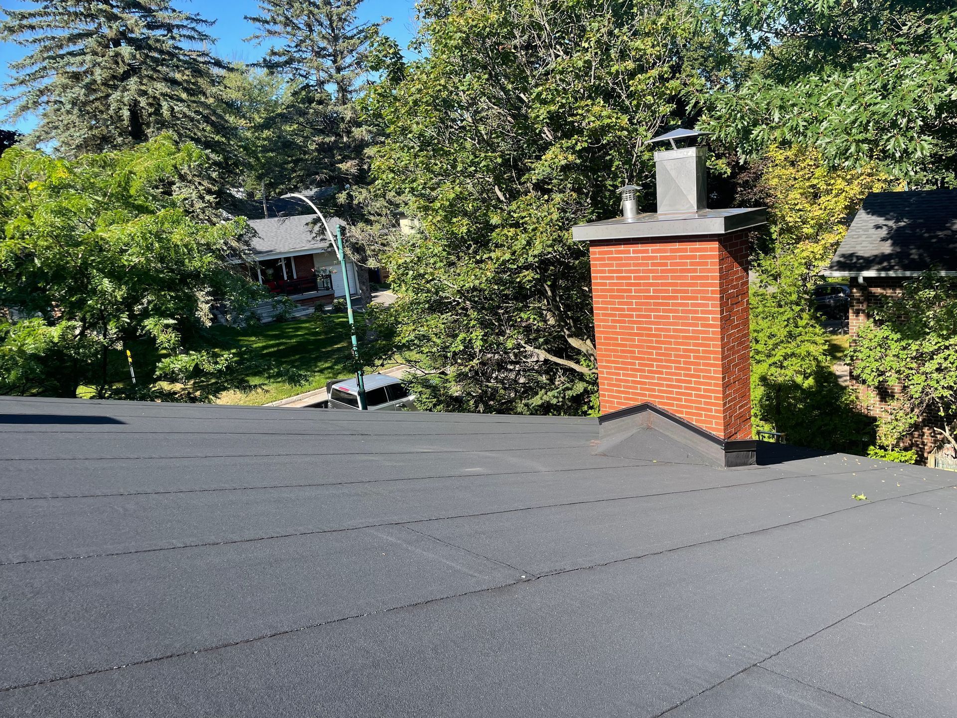 Cheminée en briques sur un toit plat noir, entourée d'arbres et d'un ciel bleu ensoleillé.