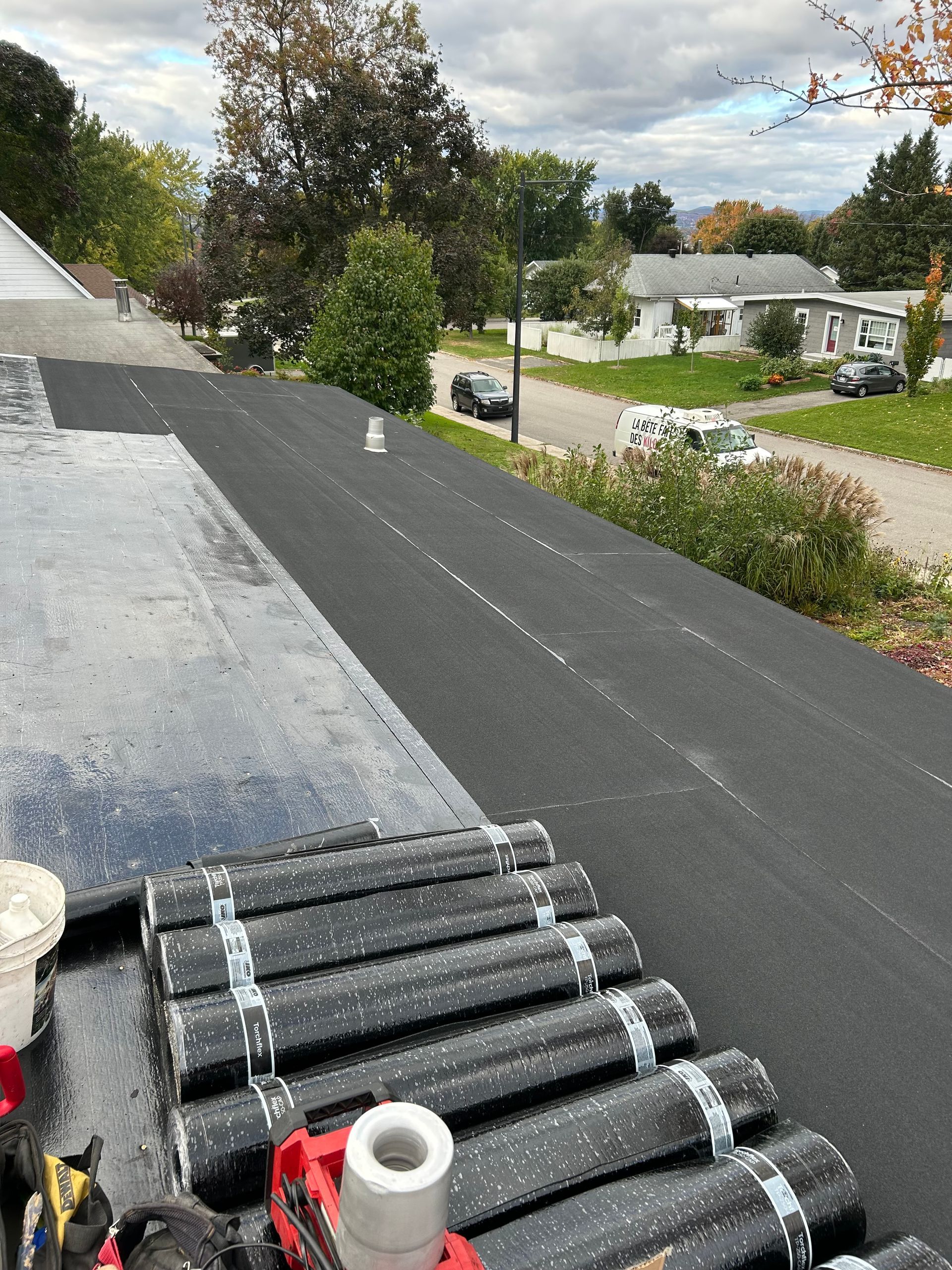 Vue du toit de la nouvelle installation de bardeaux d'asphalte; rouleaux de bardeaux, outils et un quartier de banlieue en arrière-plan.