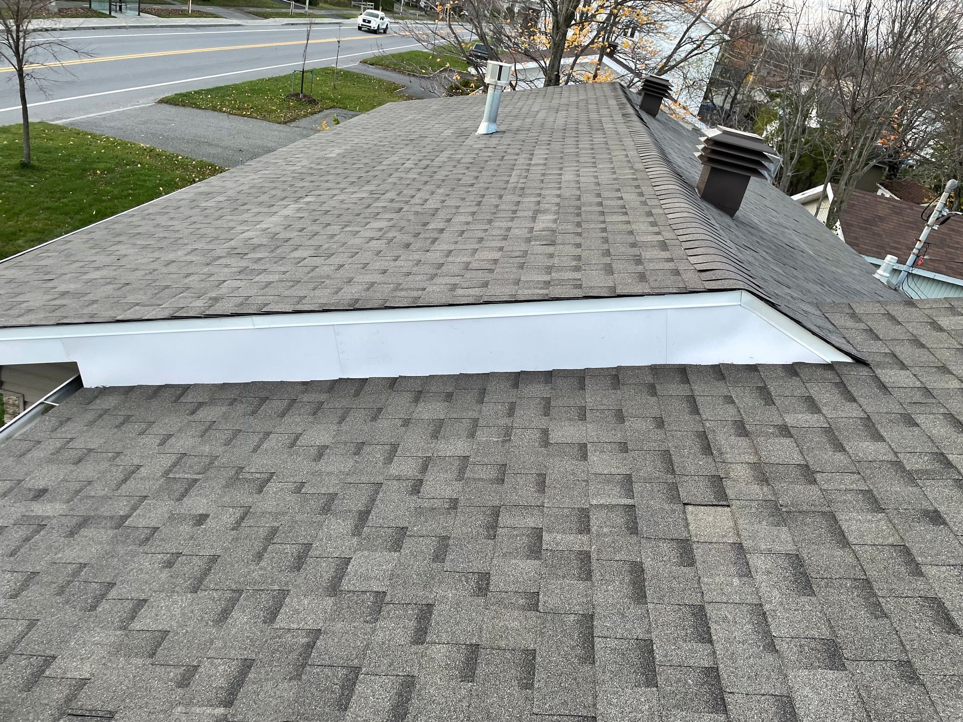 Toiture en bardeaux d'asphalte gris avec garniture blanche et cheminée, rue et herbe en arrière-plan.