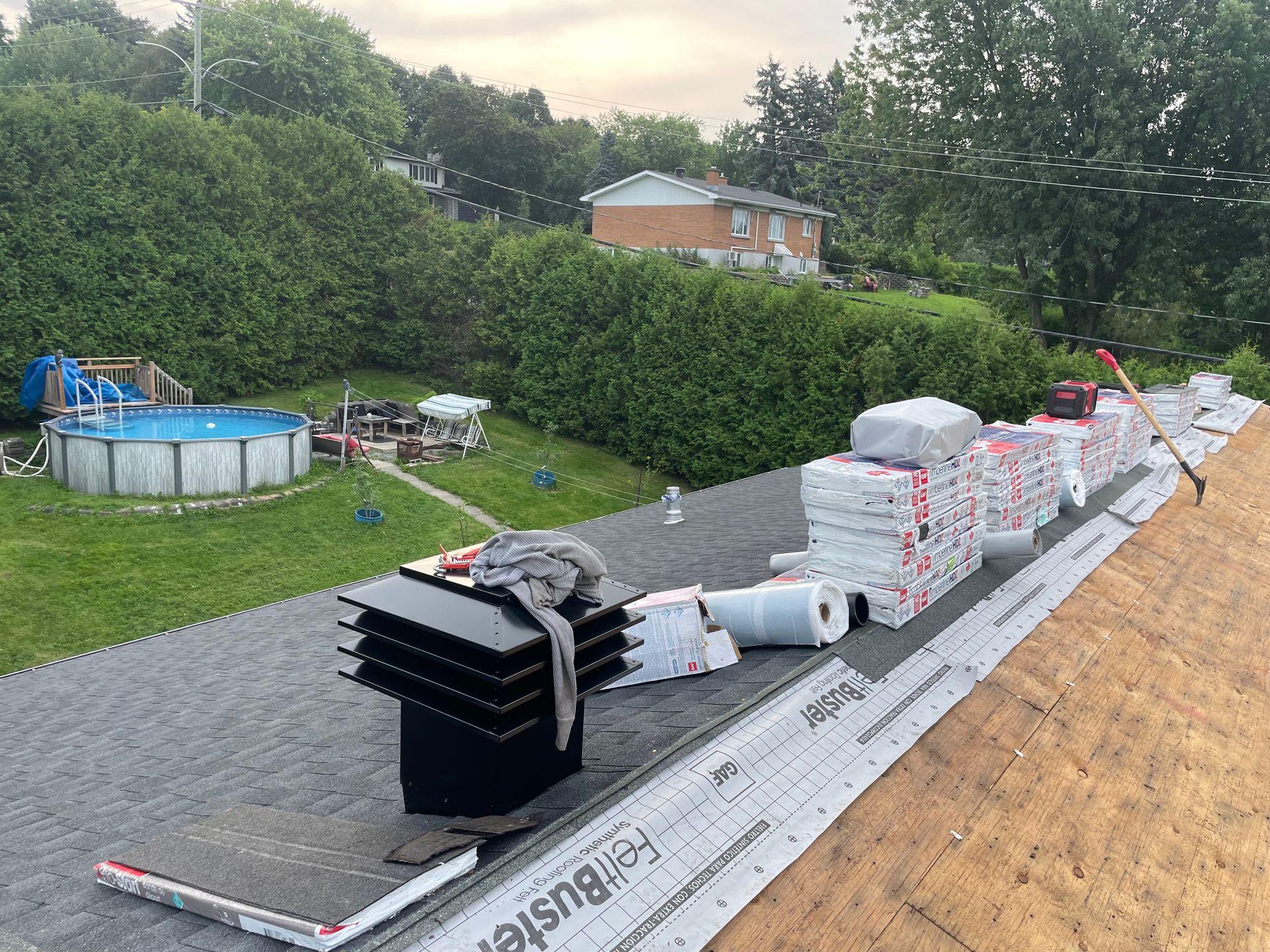 Toit recouvert de bardeaux, de matériaux de toiture et d'outils. Des maisons et une piscine sont visibles en arrière-plan.