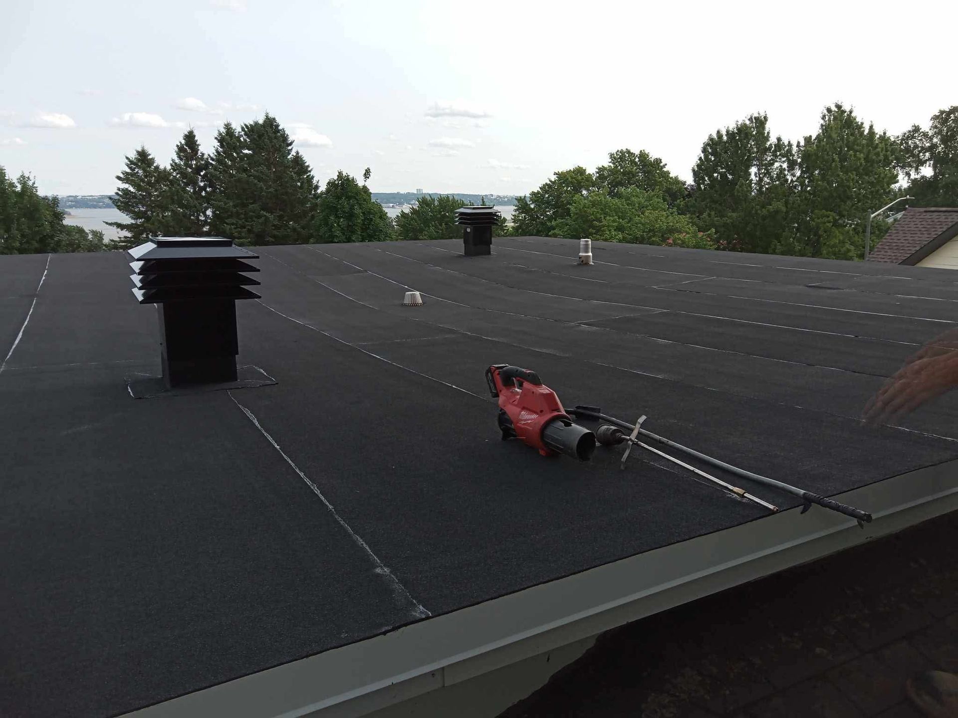 Toit plat noir avec évents; outil et câble rouges passant à la surface, arbres et eau en arrière-plan.
