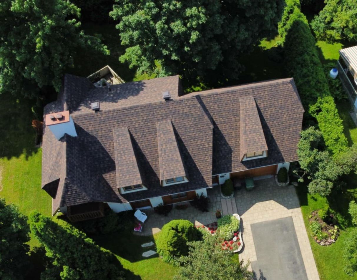 Vue aérienne d'une maison avec un toit brun, des murs blancs et une allée pavée entourée d'arbres et de verdure.