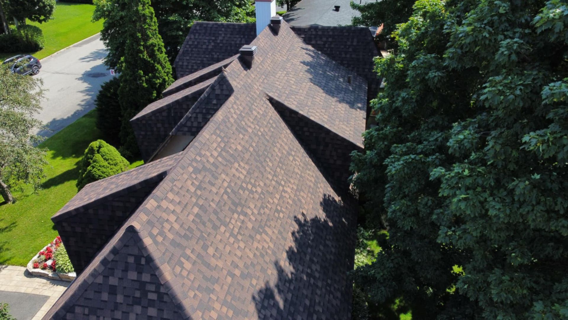 Toit en bardeaux bruns d'une maison avec une cheminée, vu d'en haut, entouré d'arbres et de verdure.