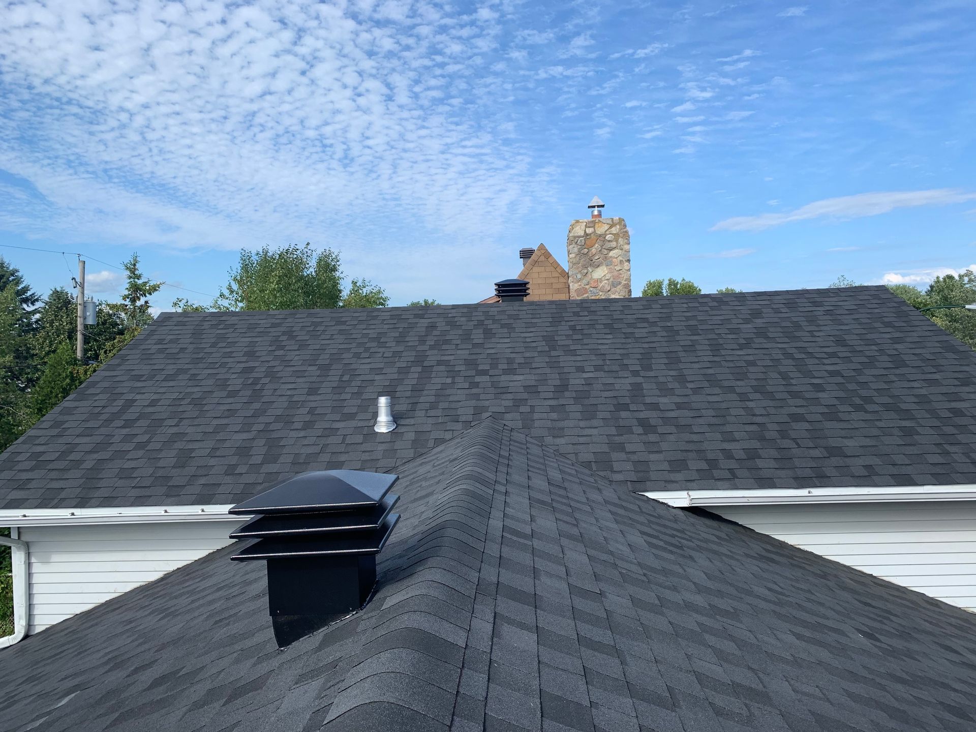 Toit en bardeaux foncés avec cheminée, gouttières blanches et ciel bleu avec des nuages.