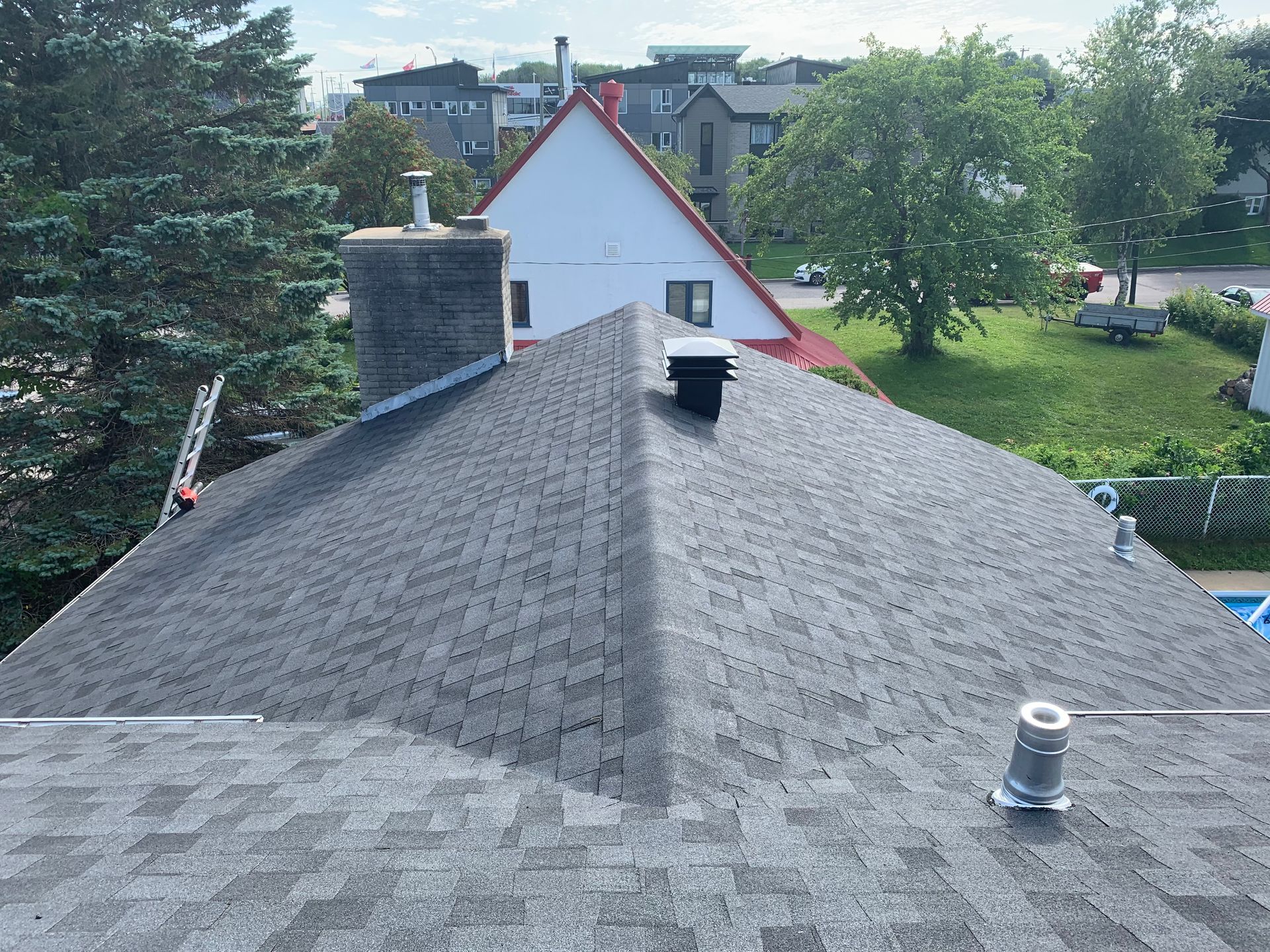 Toit en bardeaux gris avec cheminée et évents, vue sur l'arrière-cour, arbres et bâtiments en arrière-plan.