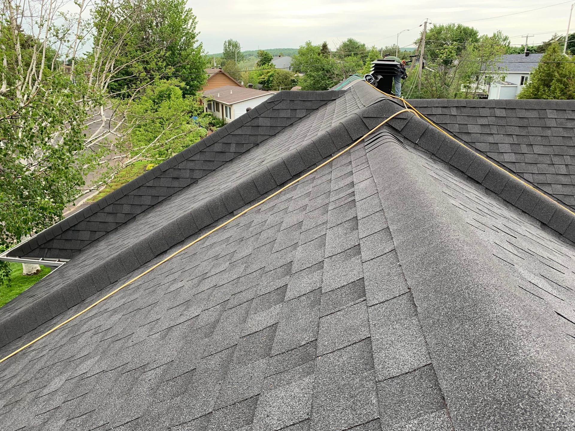 Toit en bardeaux gris foncé avec une ligne de faîte, une cheminée et une vue sur les arbres et les maisons.