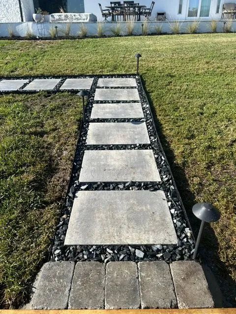 Stone path with rectangular pavers and black rock border, in a grassy backyard.