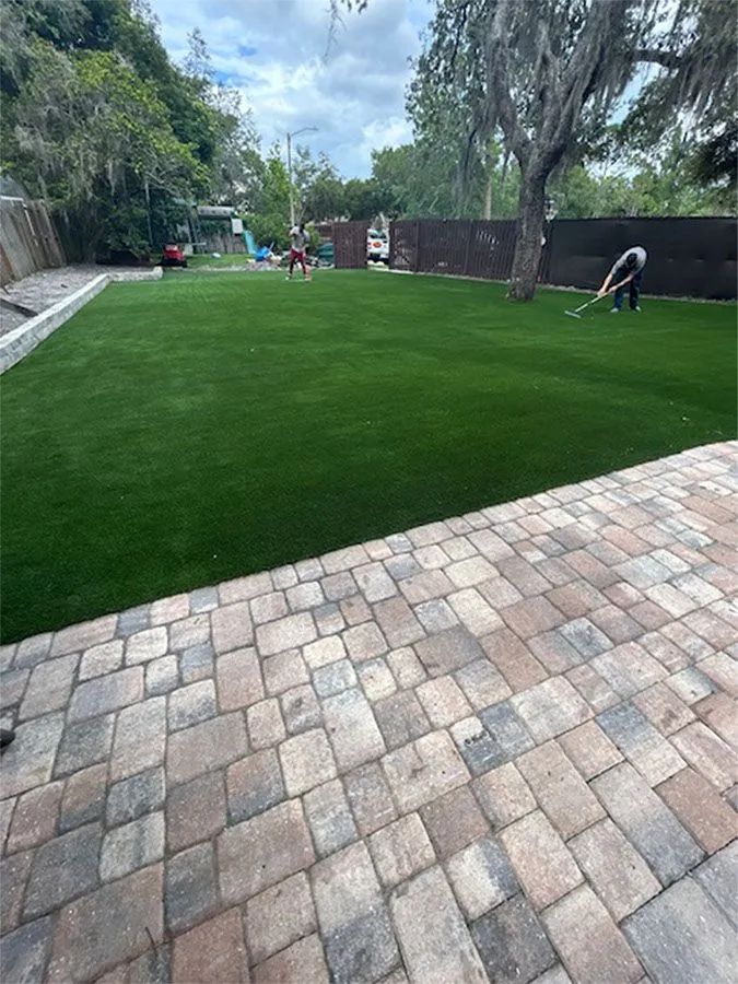 A backyard with a stone patio, vibrant green lawn, and people near a tree and fence.