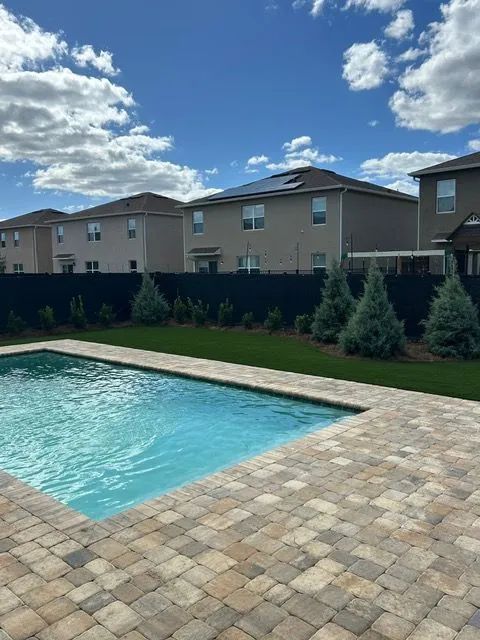 Backyard pool with paver patio, blue water, and row of houses behind a dark fence under a cloudy sky.