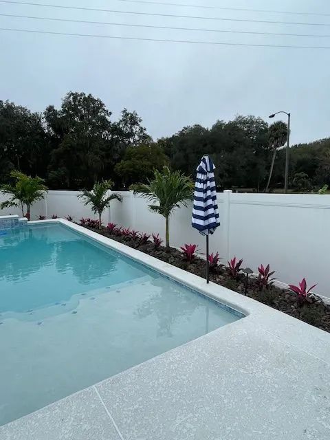 Swimming pool with a blue and white striped umbrella, bordered by plants and a white fence.