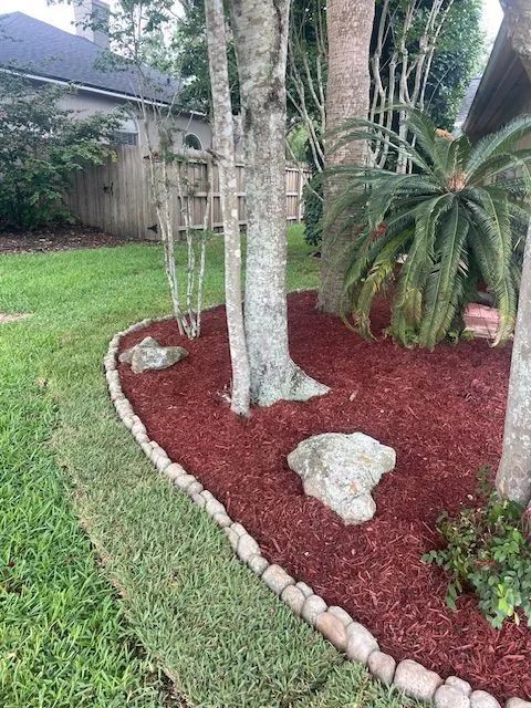 Red mulch bed with stones and trees, bordered by brown edging, next to grass.