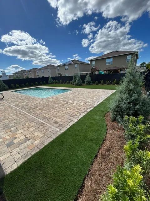 Backyard with a pool, brick patio, green lawn, and houses under a blue sky with clouds.