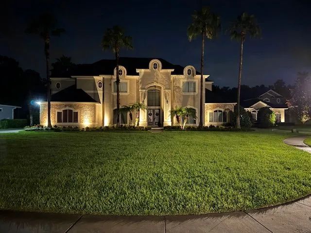 Night view of a large house with outdoor lighting illuminating the facade and front lawn; palm trees in the background.