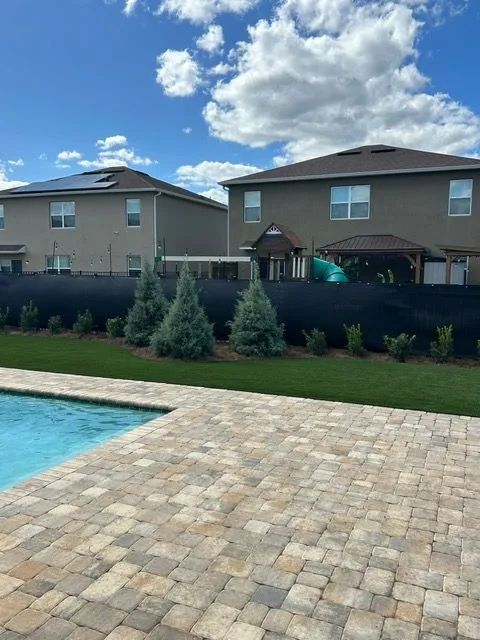 Backyard with pool, two-story houses, dark fence, blue trees, and blue sky with clouds.