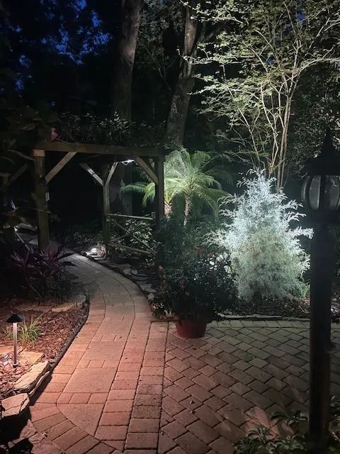 Brick pathway through a lit garden at night, passing a wooden arbor and illuminated plants.