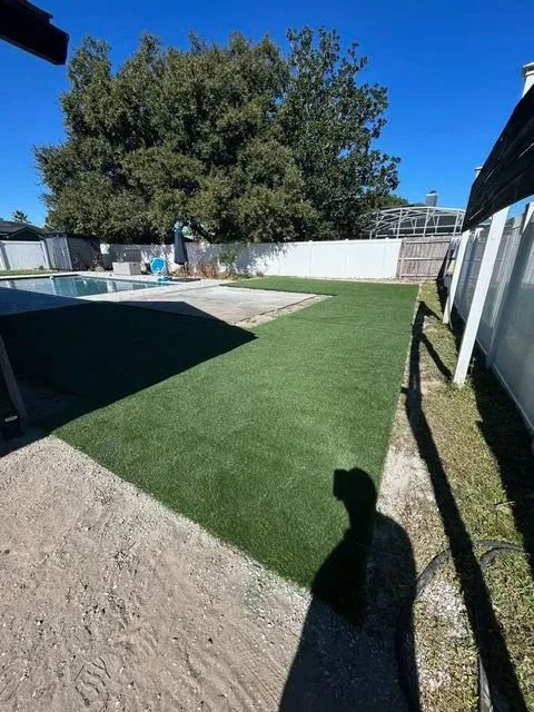 Backyard with artificial turf, concrete, a pool, and a tree under a blue sky.