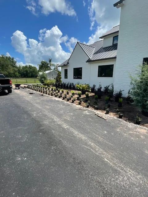 Asphalt driveway next to a white house with landscaping. Plants in pots line the border. Blue sky.